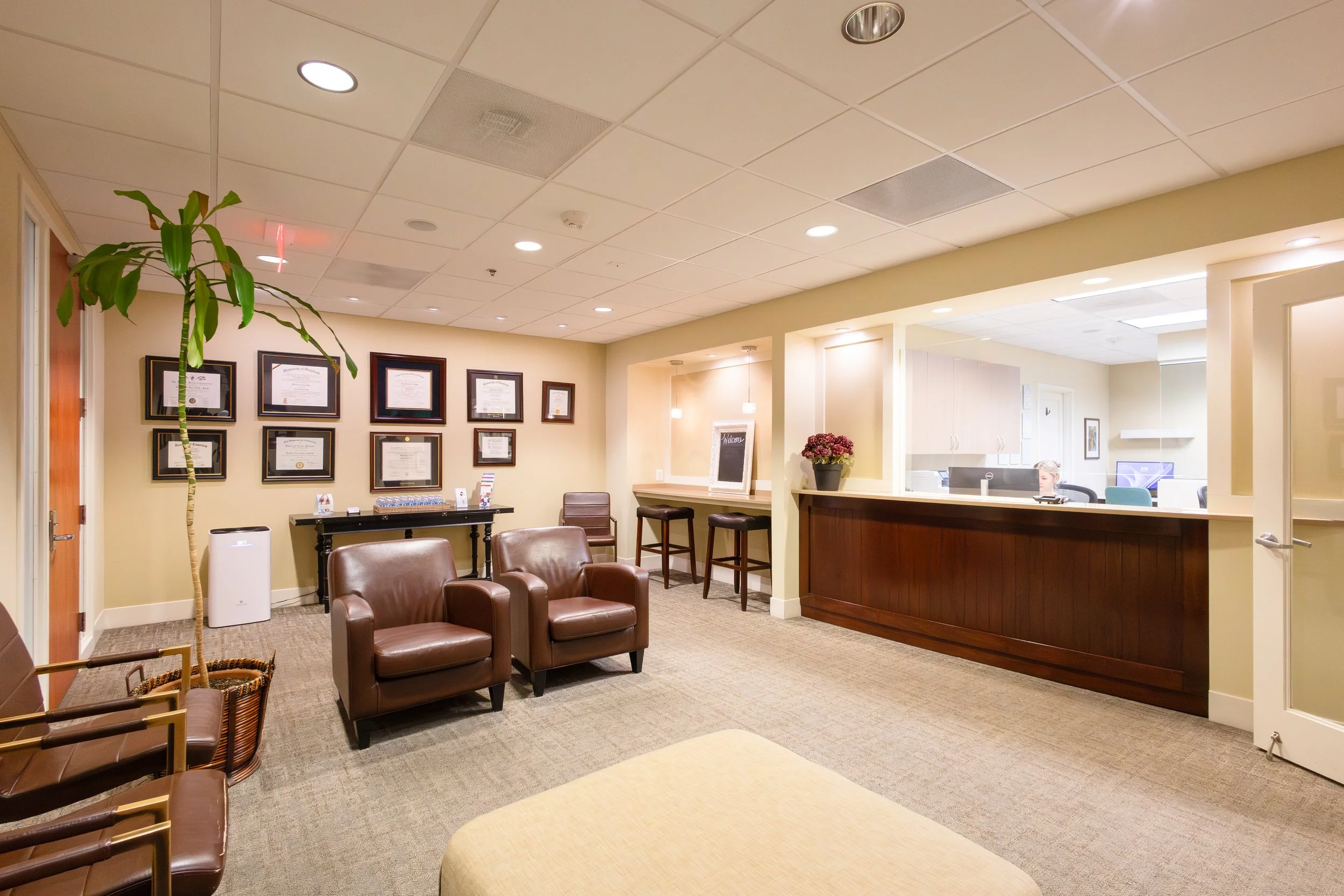 A waiting room with beige walls, brown leather chairs, a tall plant, and framed certificates on the wall. There is a reception desk with a flowerpot and a woman working behind it, with a view into a office area.