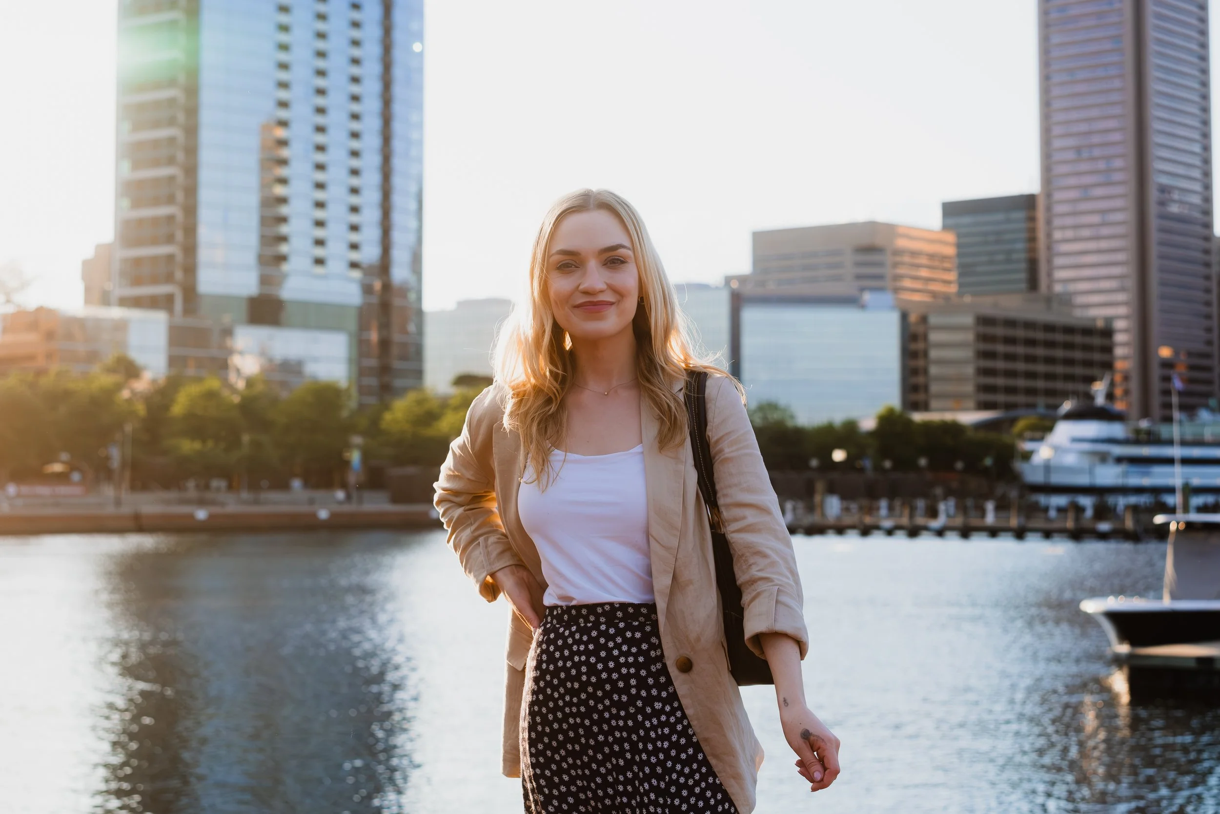 A woman standing by a river in an urban area during sunset, with tall buildings in the background.