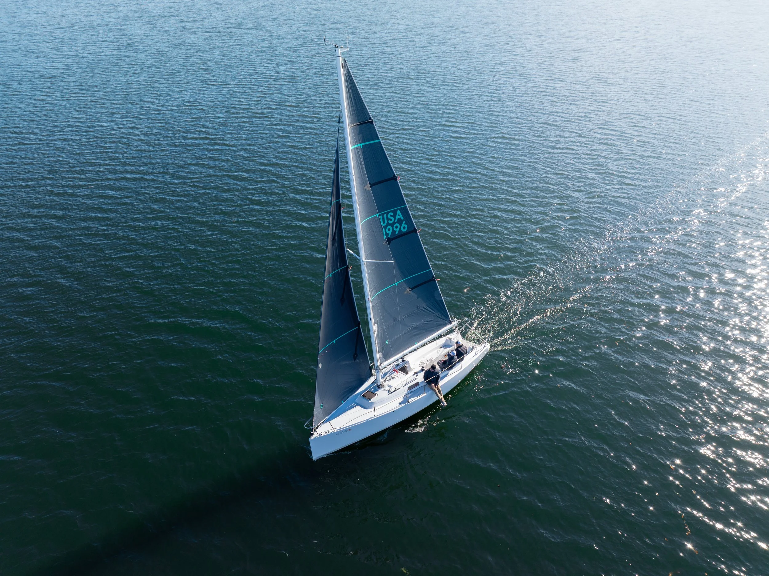An aerial view of a white sailboat with black sails marked 'USA 1996,' sailing on calm blue water with sunlight reflections.
