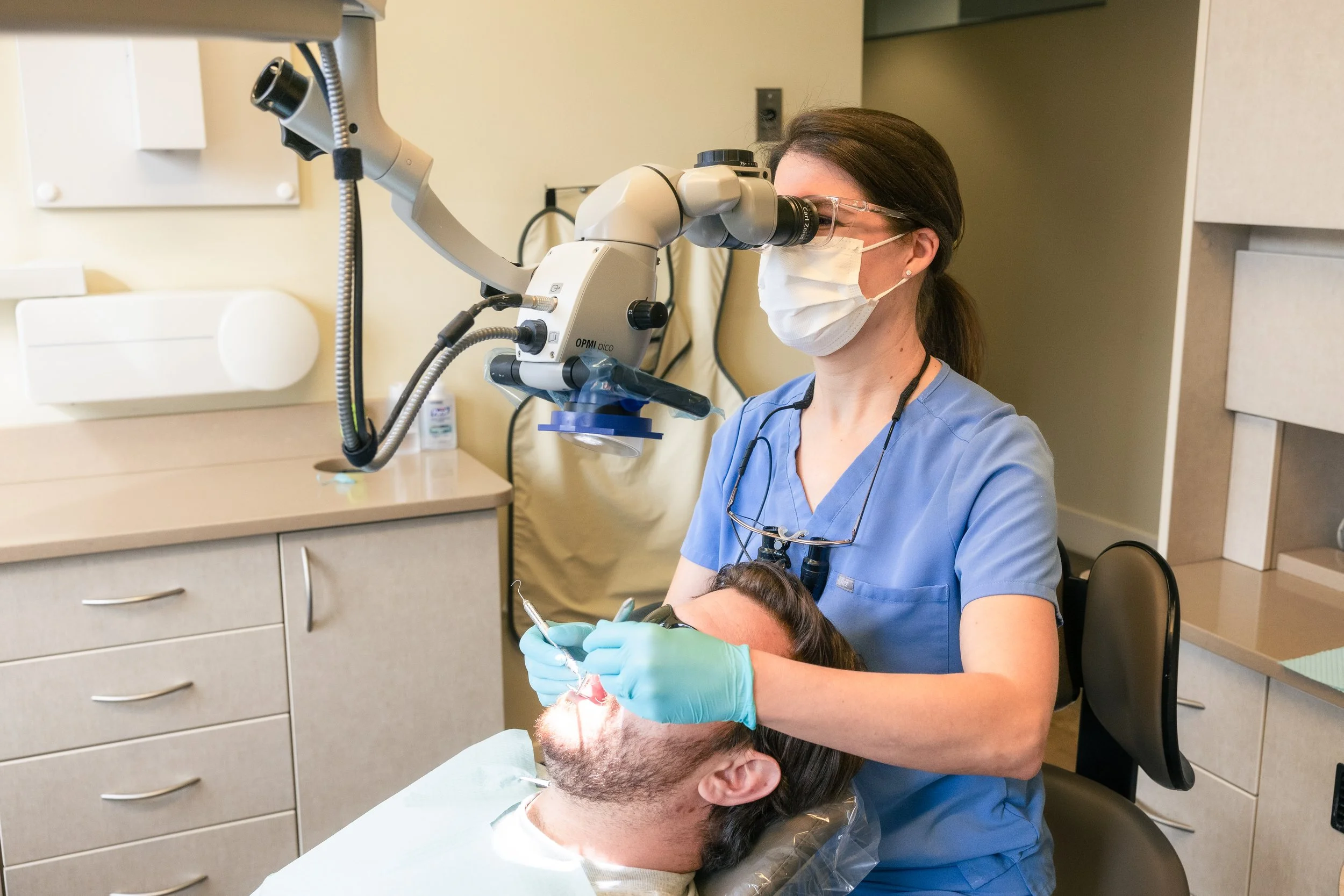 A female dentist wearing a face mask, gloves, and scrubs using a dental microscope to examine or treat a male patient lying in a dental chair.