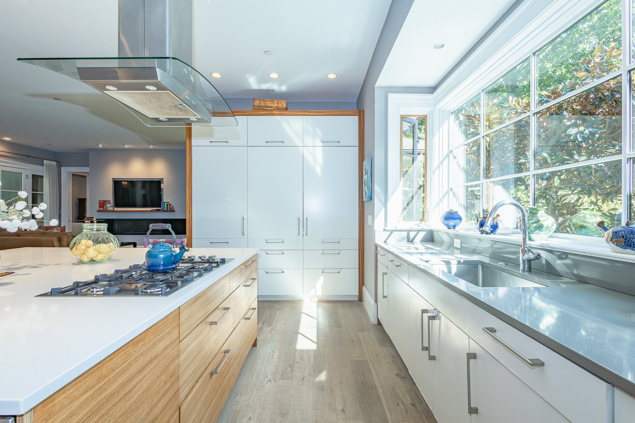 Bright kitchen with large window, white cabinetry, wooden accents, and a kitchen island with a blue teapot and glass jar on it.