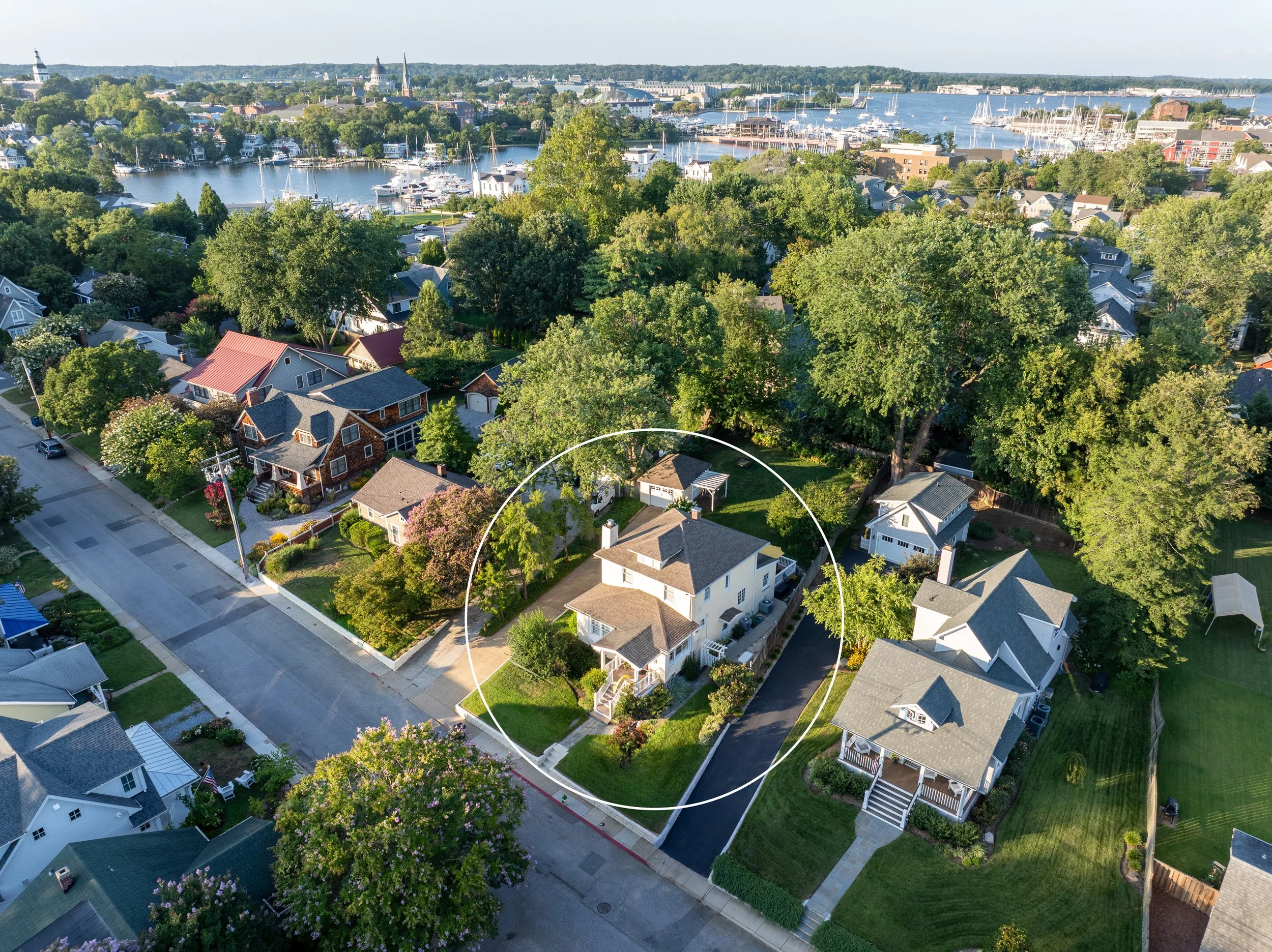 Aerial view of a neighborhood with multiple houses, trees, and a body of water with boats in the background. A specific house is outlined with a white circle.
