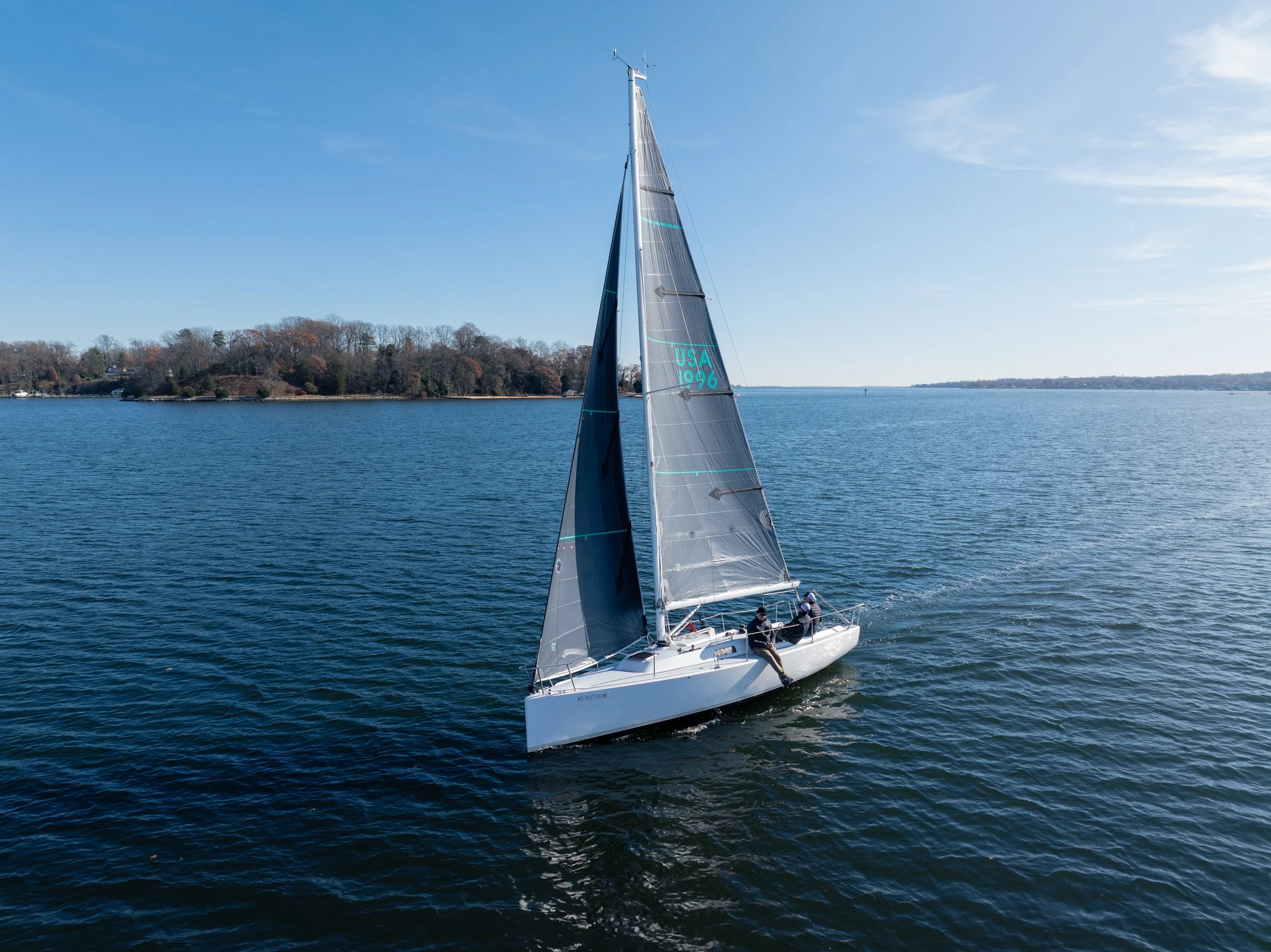 A white sailboat with a crew of three people sailing on a calm body of water, with forested land in the background and a clear blue sky.