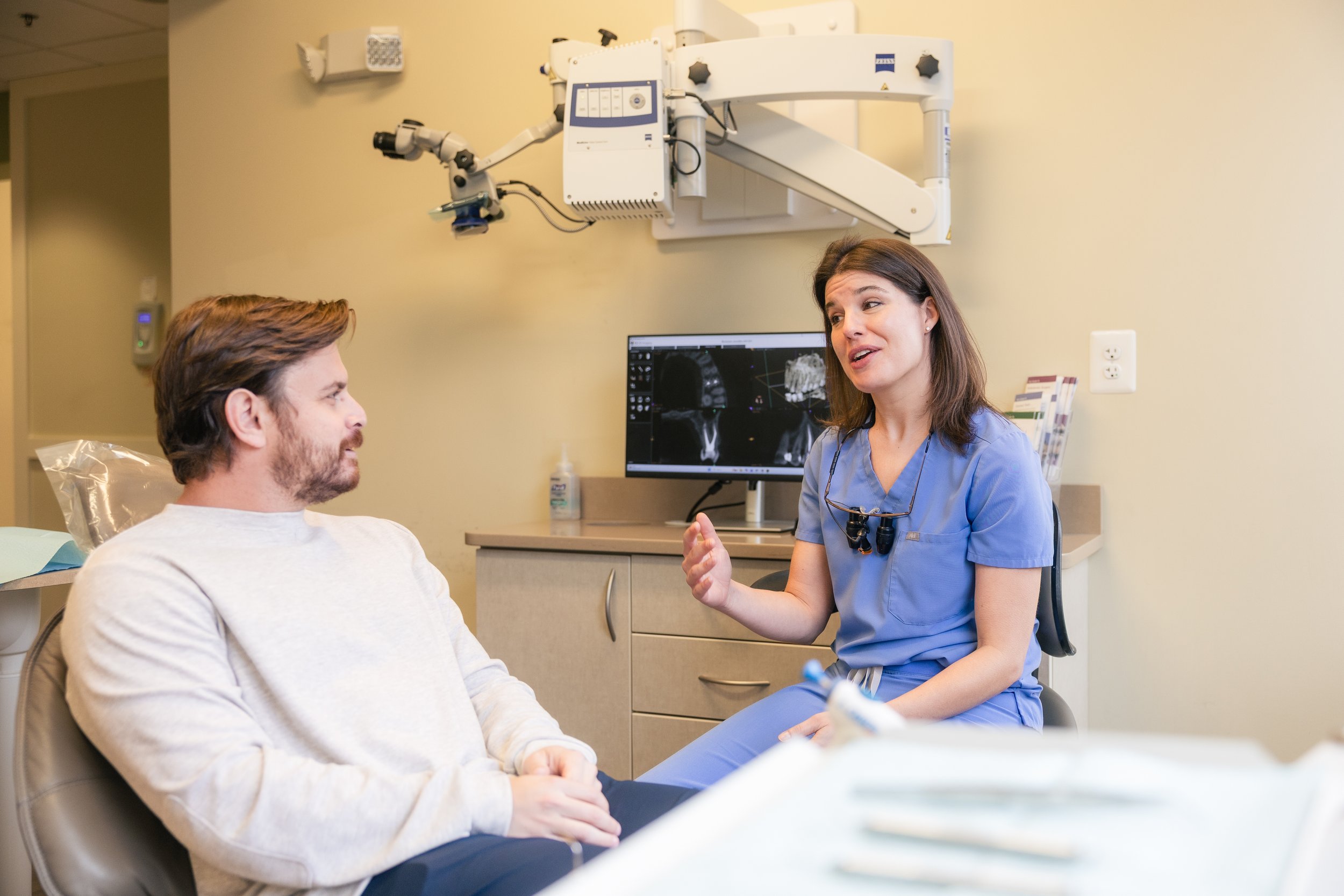 A female healthcare professional in blue scrubs talking to a male patient in a medical exam room. Behind her, a monitor displays medical scans.