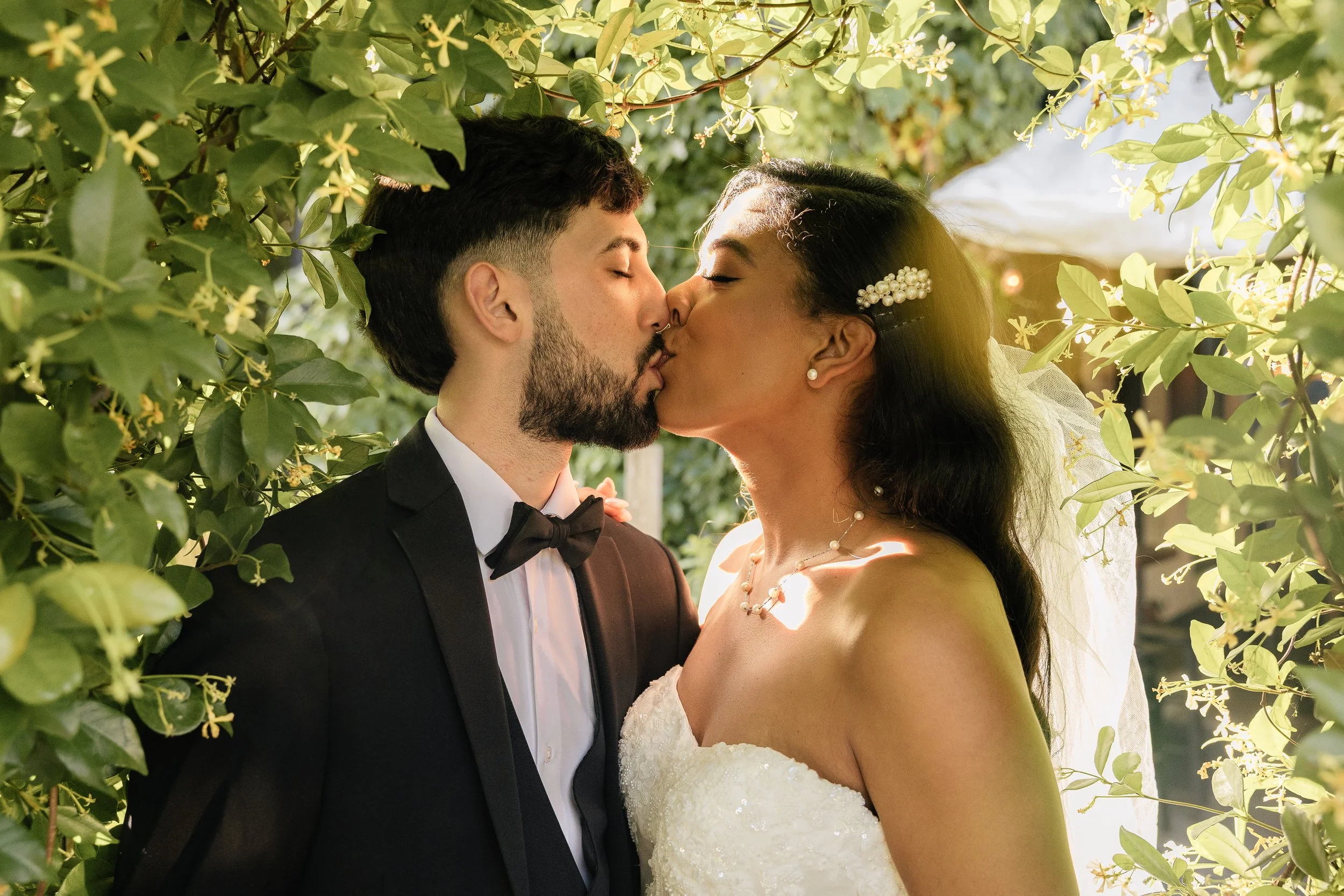 A newlywed couple shares a kiss amidst green foliage, with sunlight filtering through the leaves. The groom is dressed in a black tuxedo with a bow tie, and the bride is wearing a strapless white wedding gown adorned with pearl jewelry.