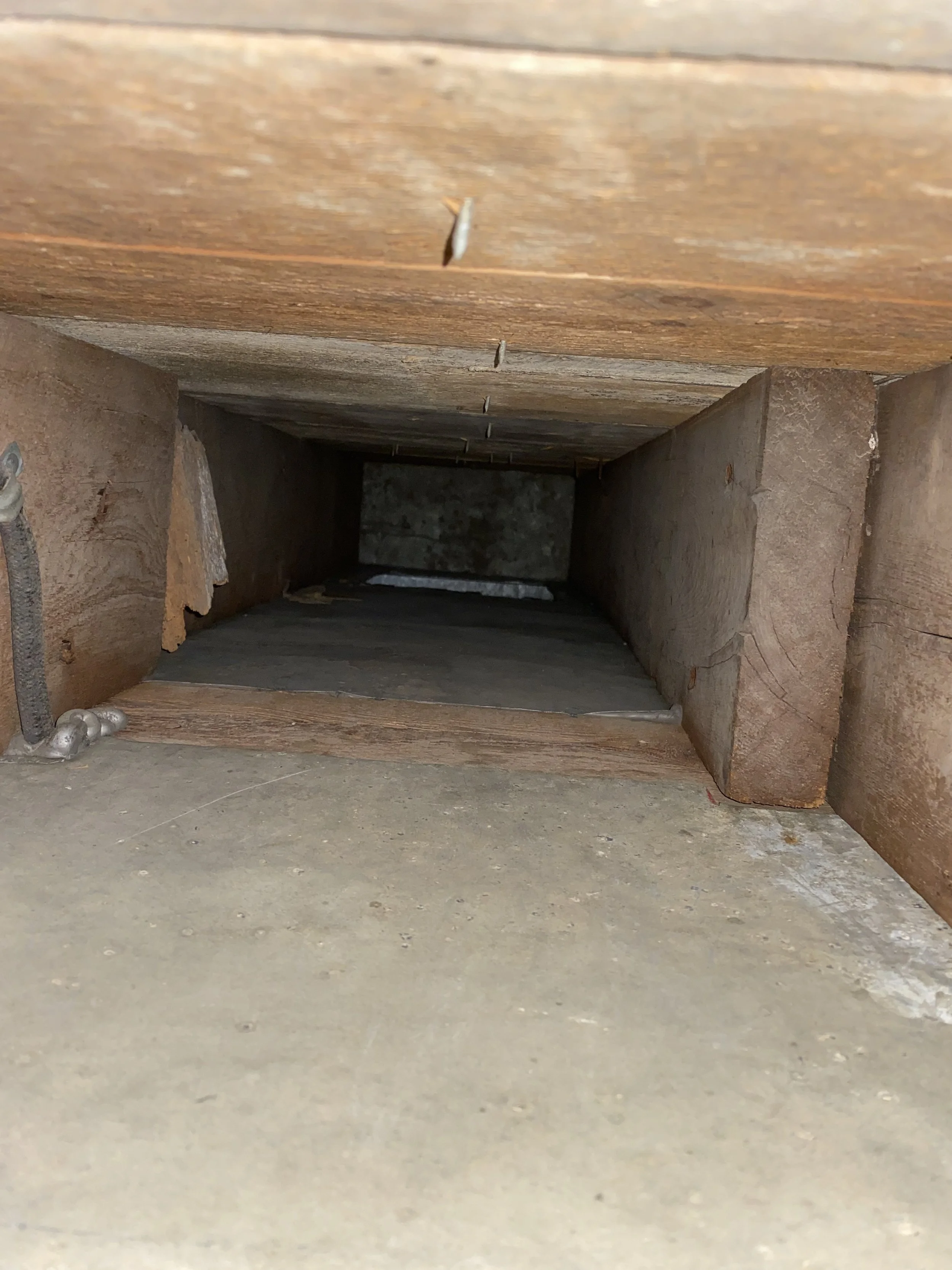 Looking into a narrow wooden and concrete crawl space or vent with visible nails, wooden boards, and concrete flooring.