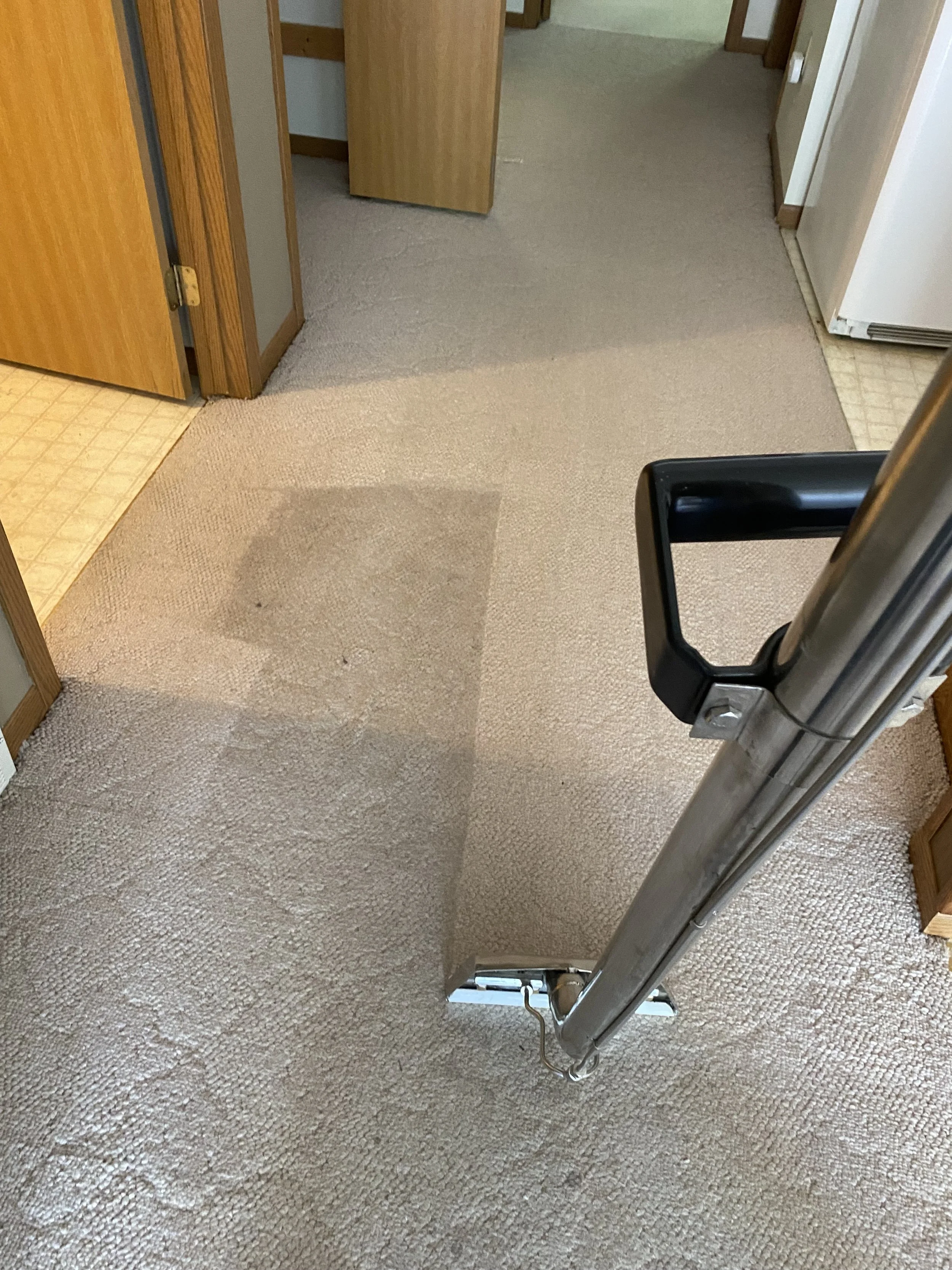 A view of a carpeted floor with a section vacuumed, revealing the cleaner underneath. Part of a metal cleaning tool with a black handle is visible in the lower right corner. The surrounding area includes a doorway, wooden cabinet, and part of a kitchen with tiled floors.