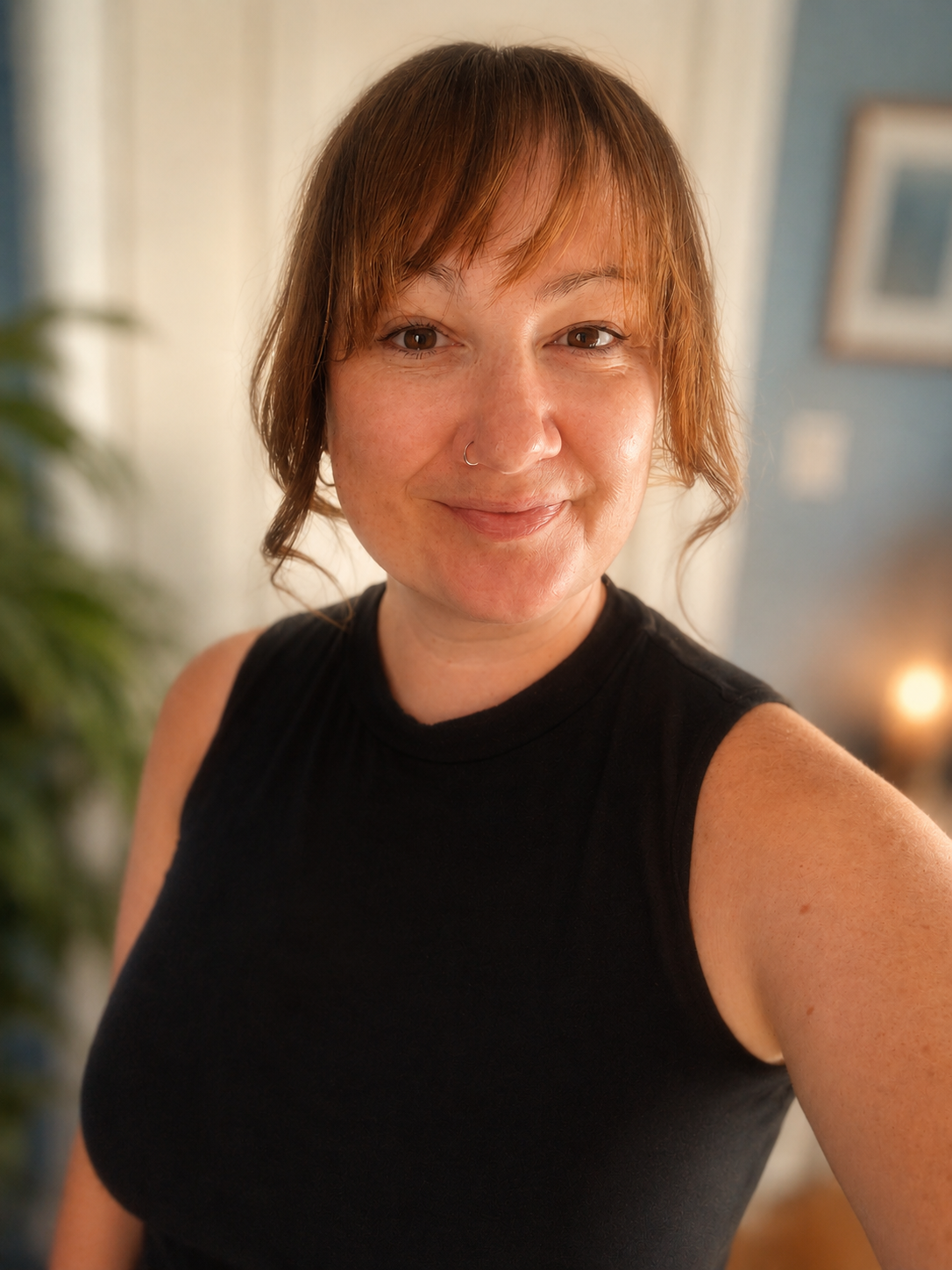 Close-up of a woman with short reddish-brown hair, wearing a black sleeveless top, smiling at the camera in a warmly lit room.