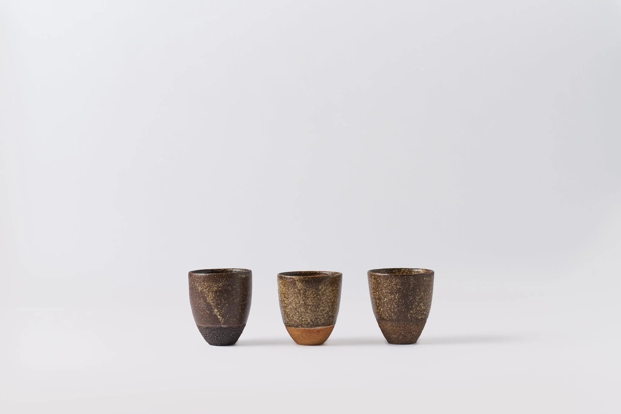 Three brown ceramic cups with textured surface and rounded shape are arranged in a row against a plain white background.