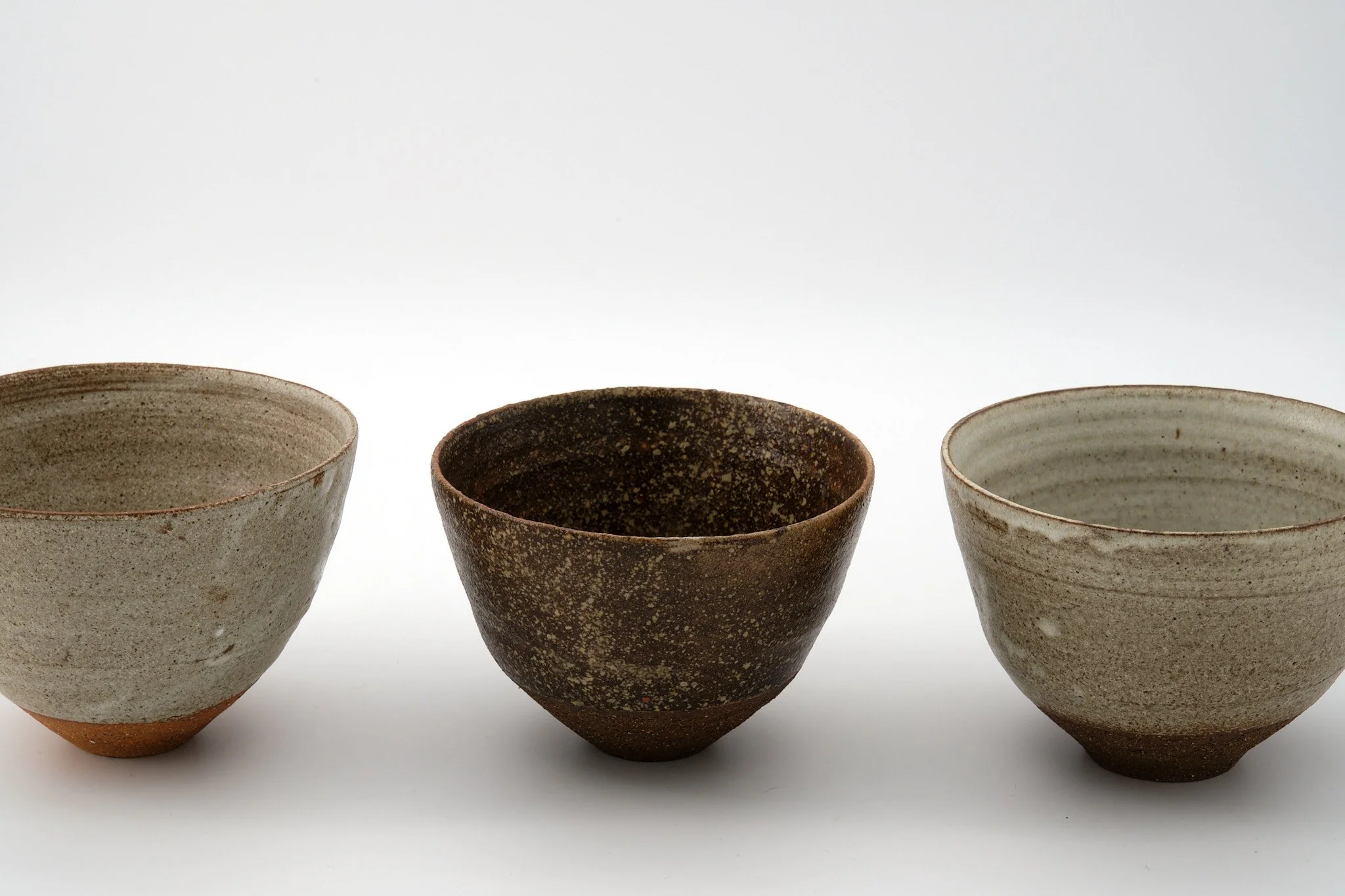 Three ceramic cups with earthy tones and speckled textures are lined up in a row against a plain white background.
