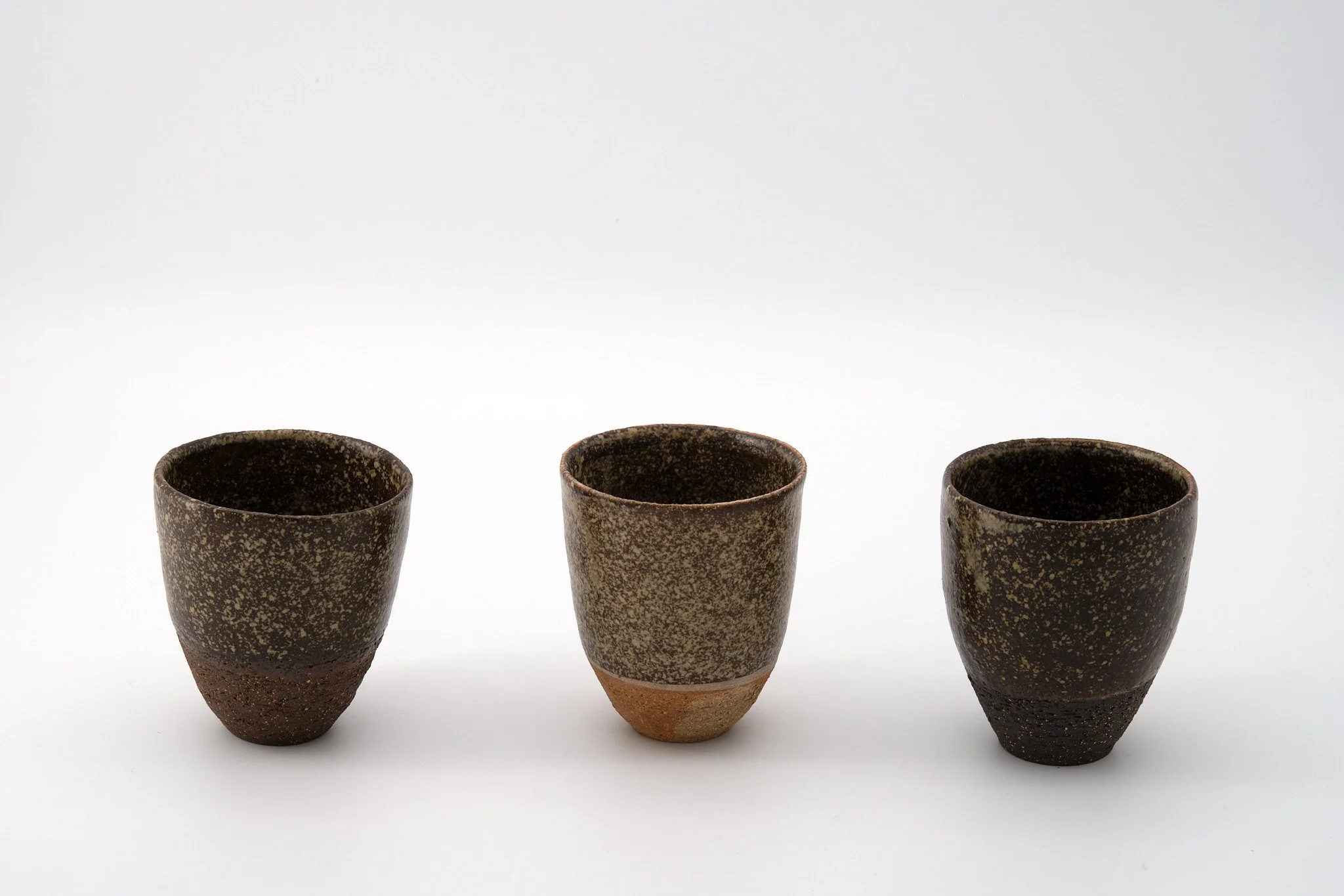 Three pottery cups with dark speckled glaze on a white background.