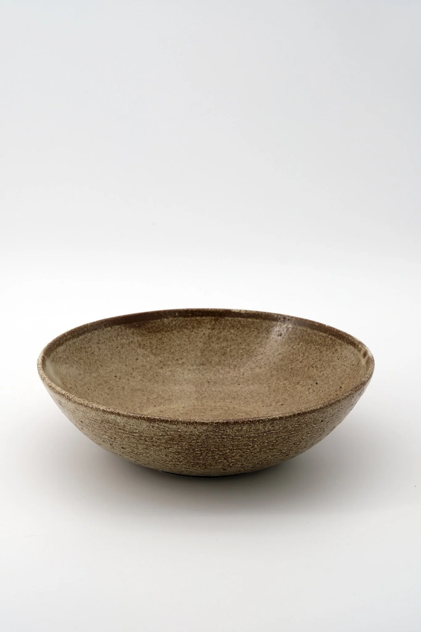 A round, textured ceramic bowl with a rough, speckled brown exterior and beige interior placed on a white surface against a white background.