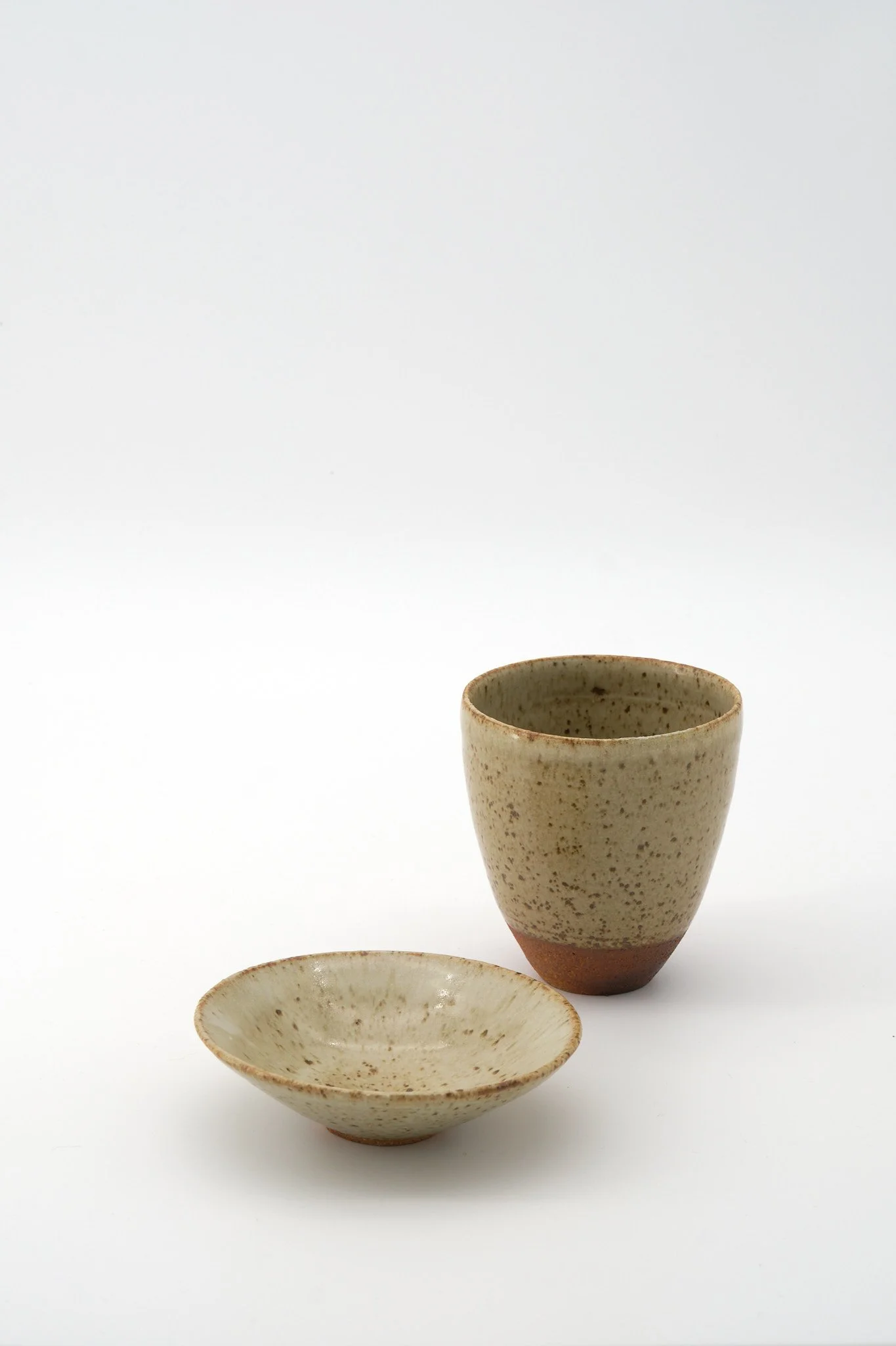 A set of three handmade ceramic tableware pieces: a tall beige speckled cup, a matching smaller bowl, and a flat shallow dish, all arranged against a white background.