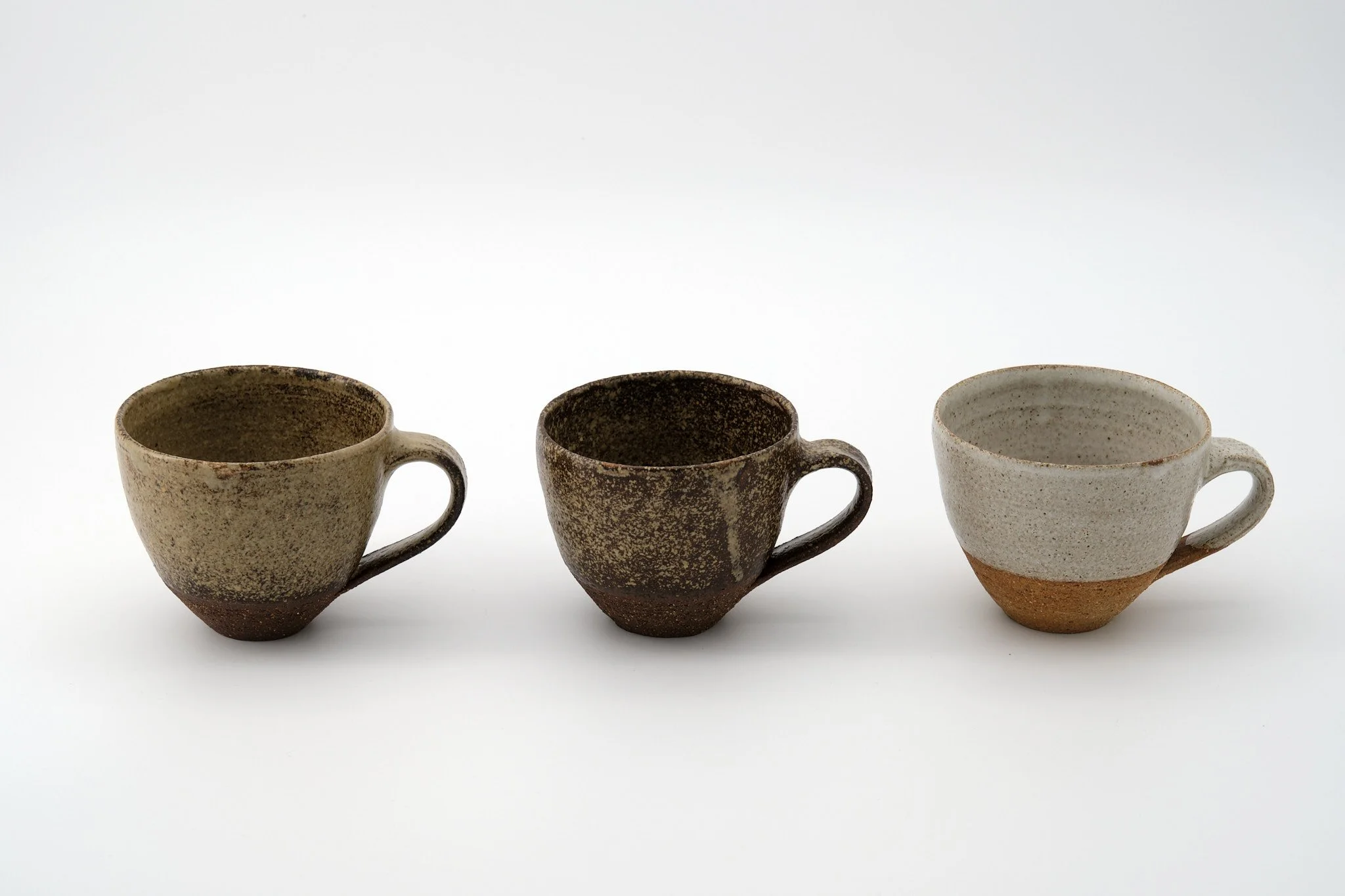 Three ceramic mugs with textured, earthy glaze and handles, arranged in a row on a white background.