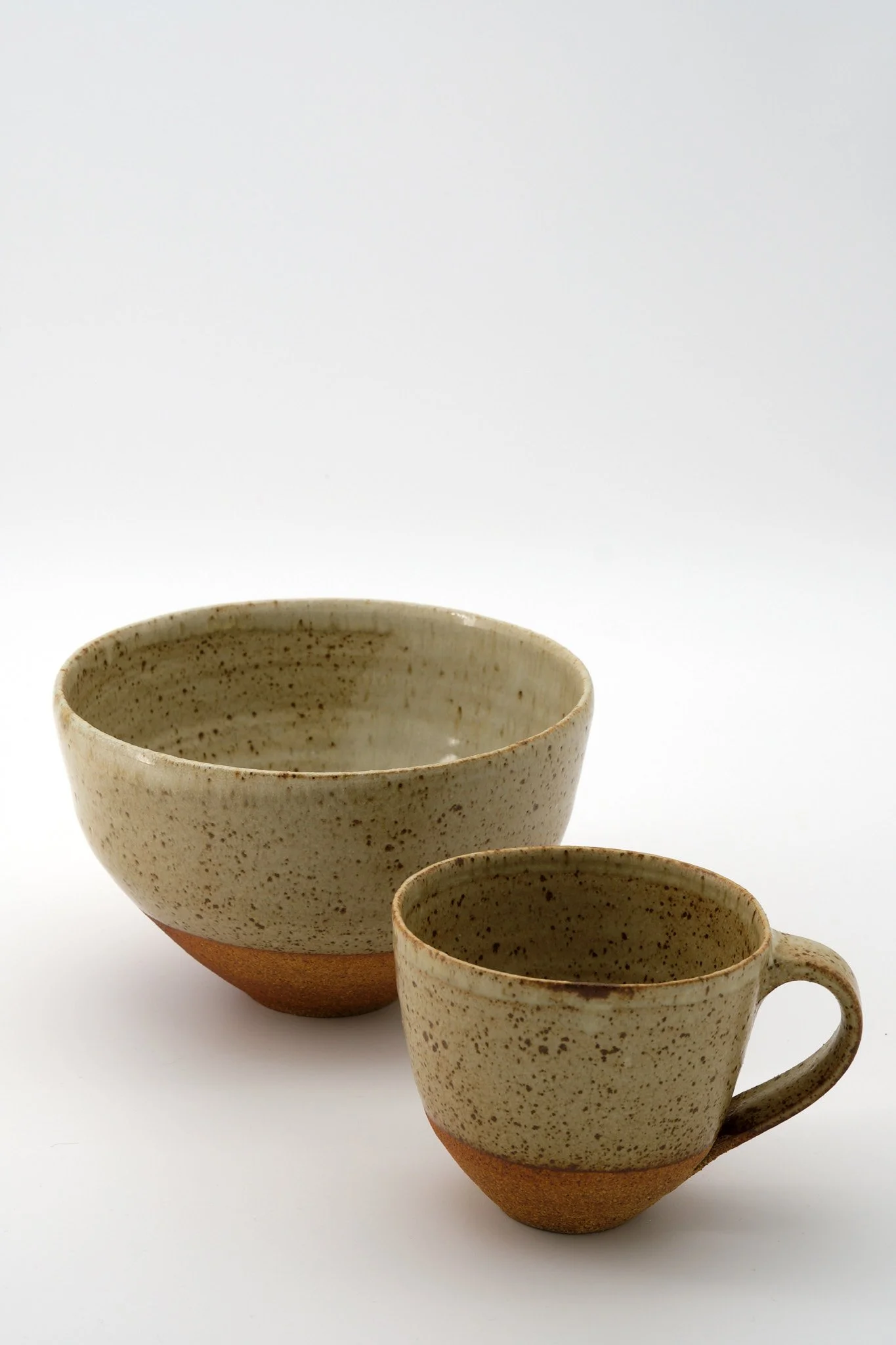 Two ceramic cups with speckled beige glaze and a reddish-brown base, set against a plain white background.