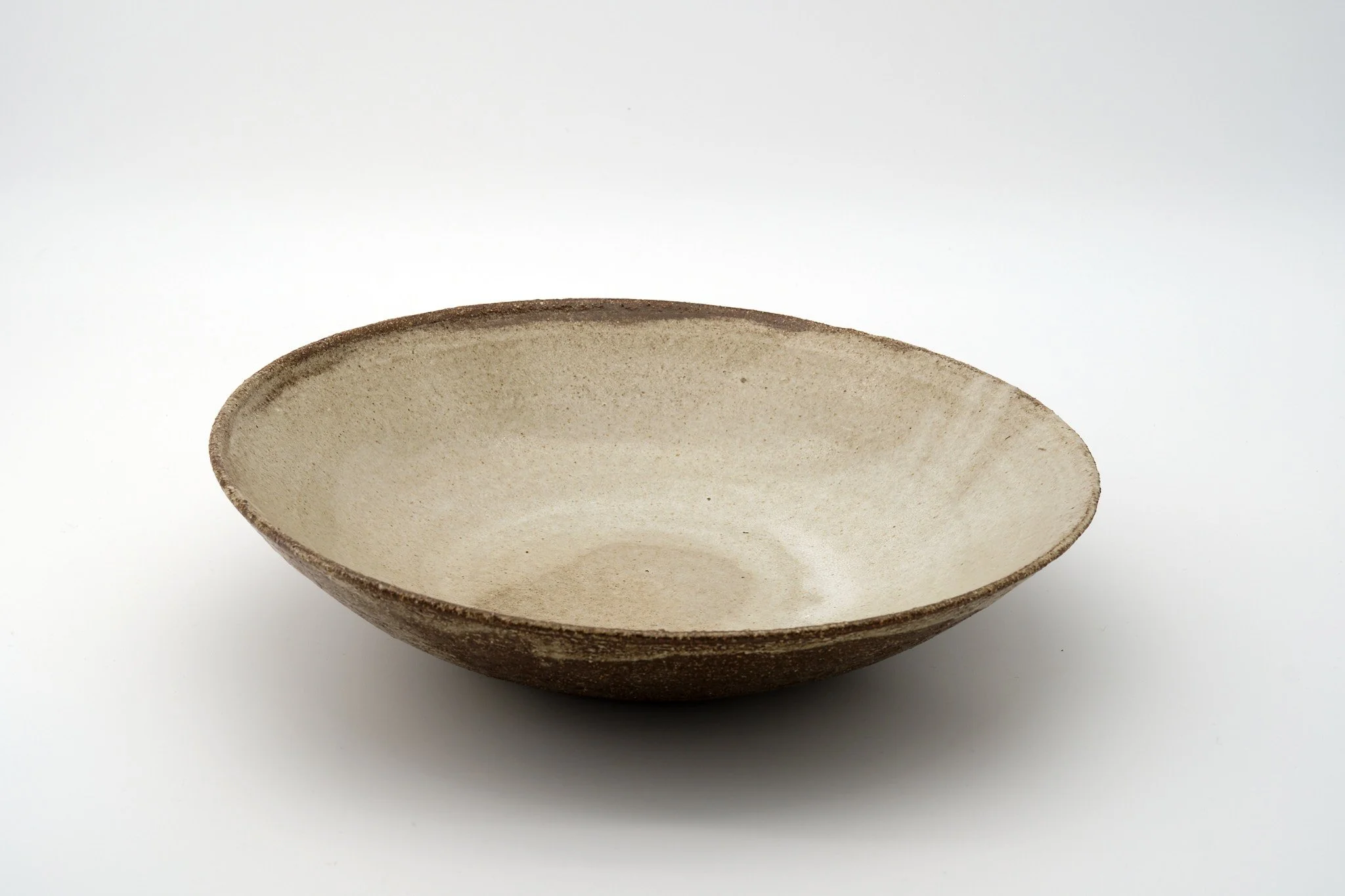 A shallow, round ceramic bowl with a rough, textured exterior and a smooth, light-colored interior, placed on a white surface.