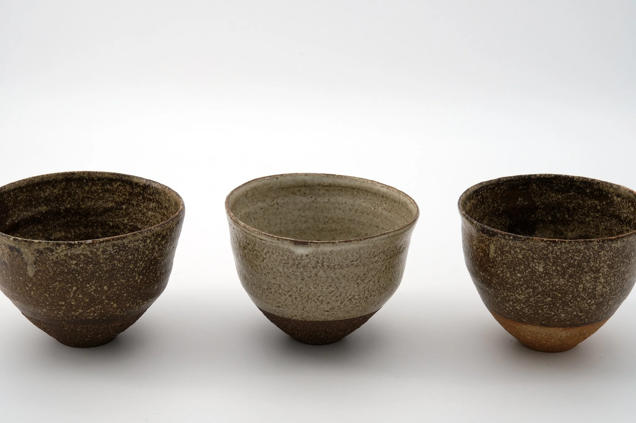 Three ceramic cups with a speckled, earthy glaze, displayed in a row against a plain white background.