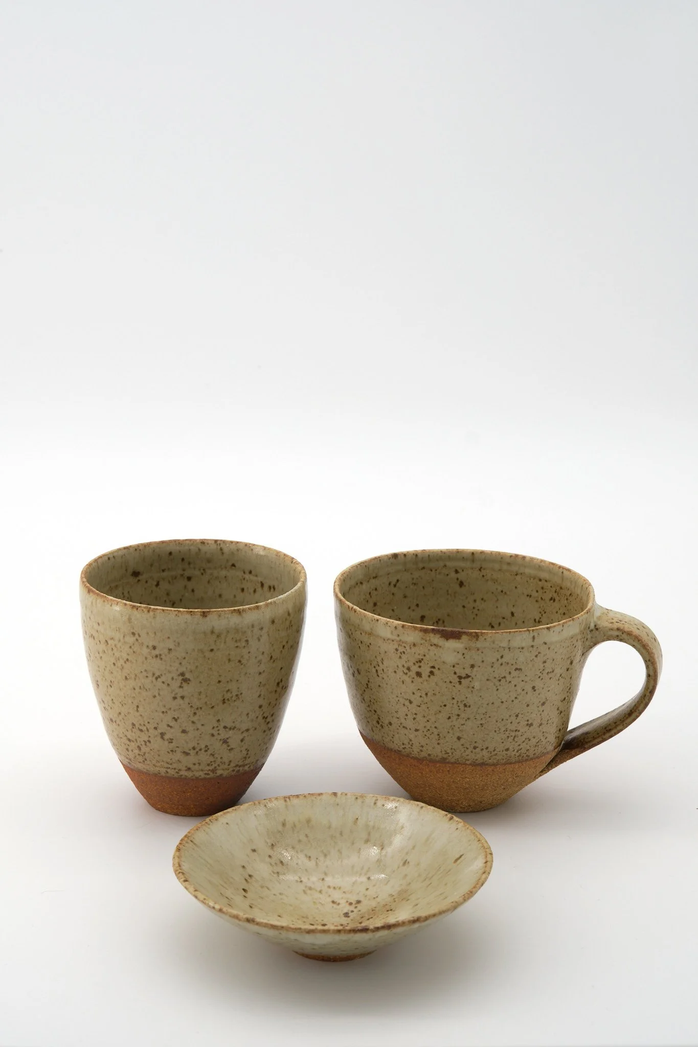 Three speckled beige ceramic cups with one matching bowl in front, all against a white background.