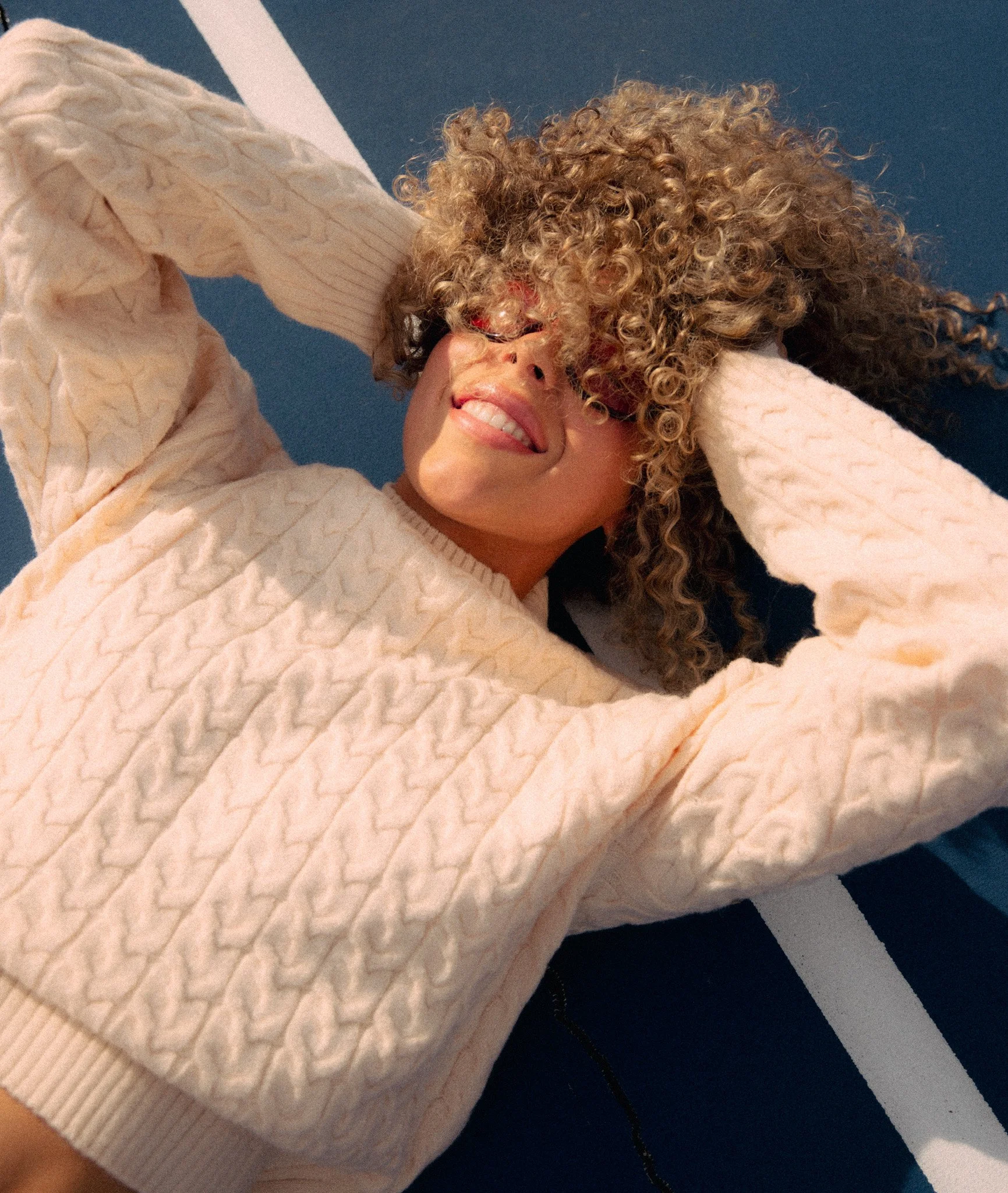 Woman with voluminous curly hair and red-tinted sunglasses smiles while lying on a blue sports court in a cream cable knit sweater, lit by golden sunlight.