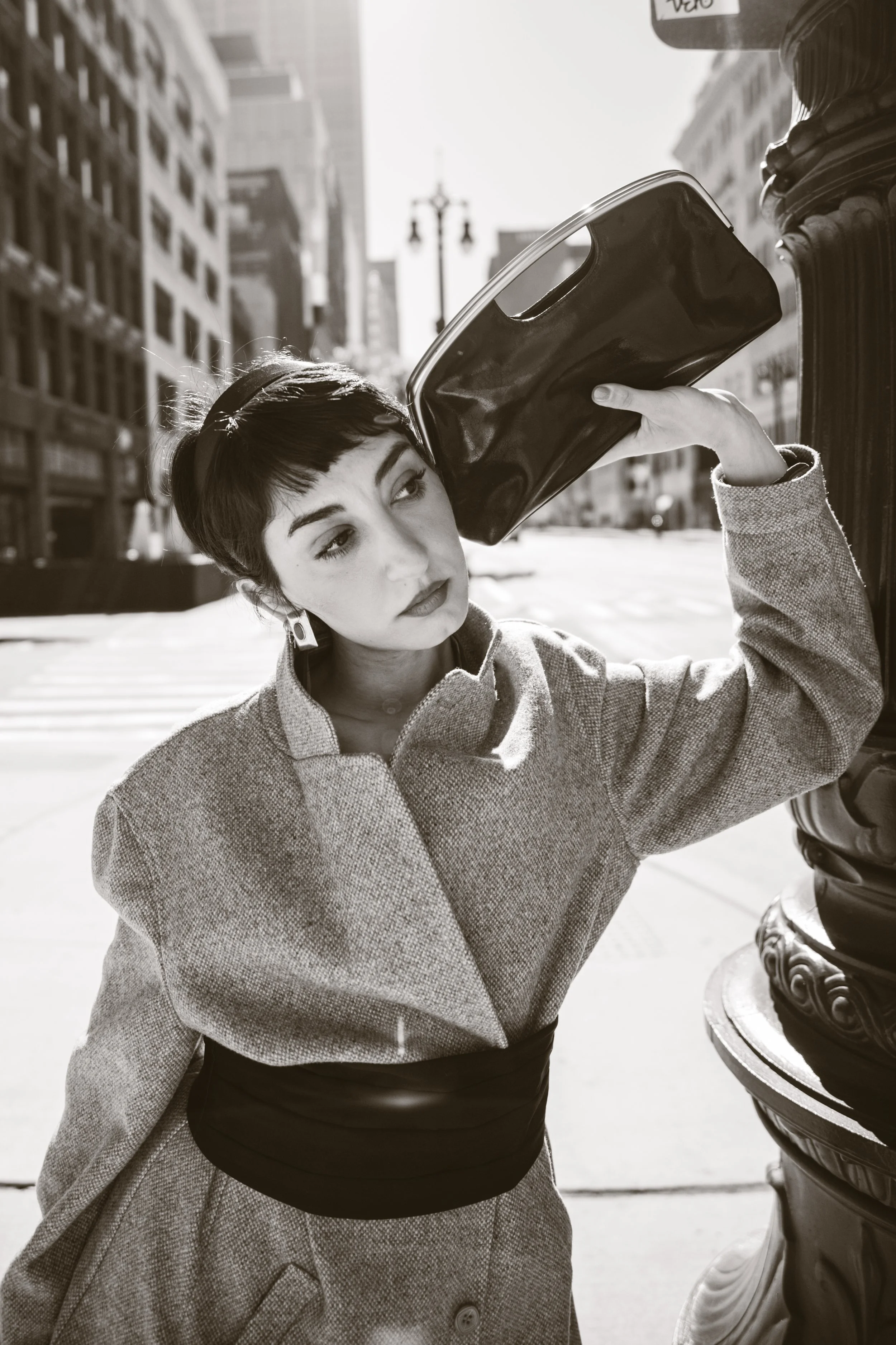 Black and white editorial fashion portrait of a model in a tweed coat holding a patent leather clutch against a lamppost in downtown Detroit, Michigan, by Olivia Feldman Photography