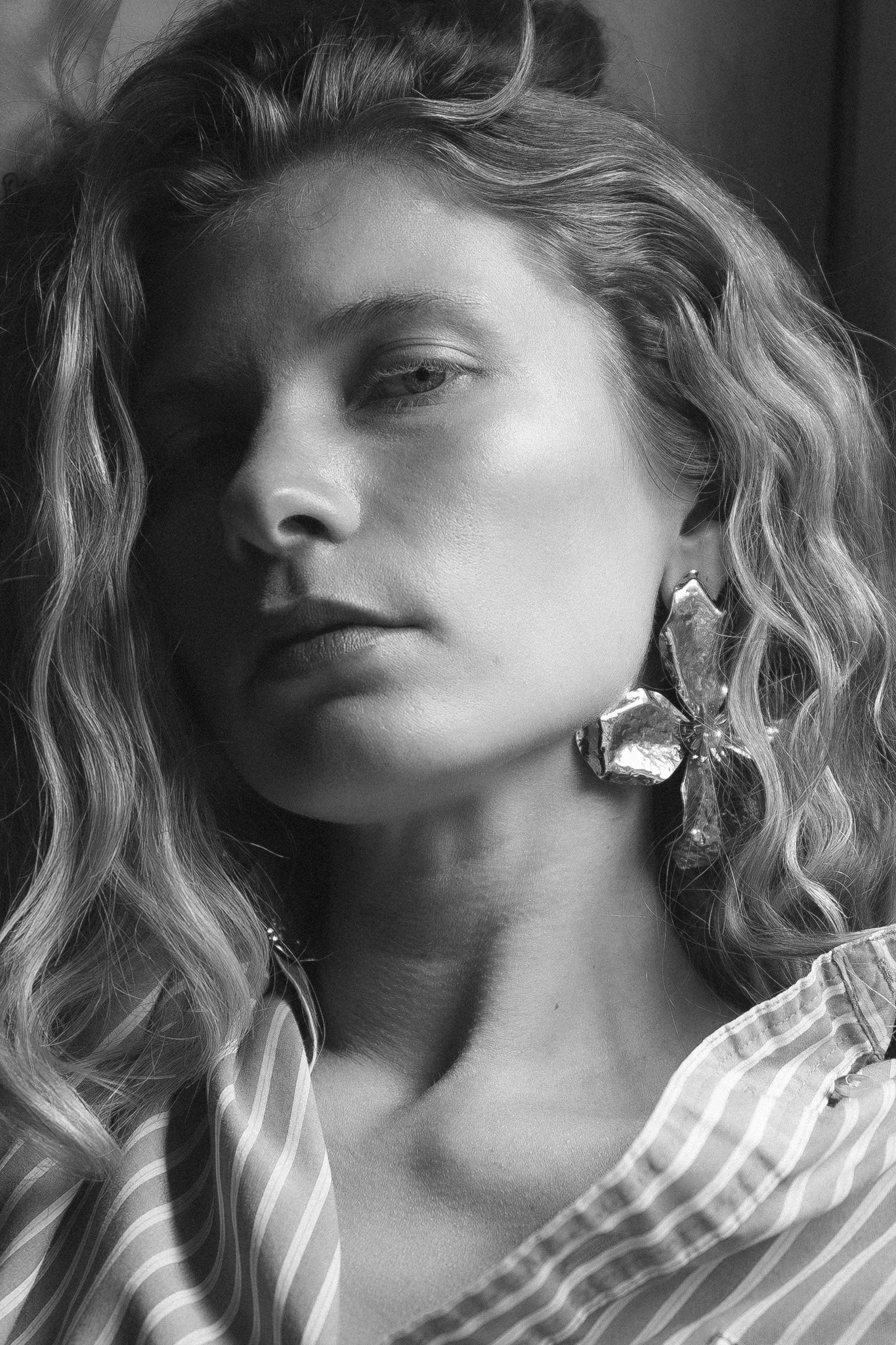 Black and white close-up portrait of a woman with wavy hair gazing to the side, wearing a large sculptural flower earring and a striped button-up shirt in soft window light.