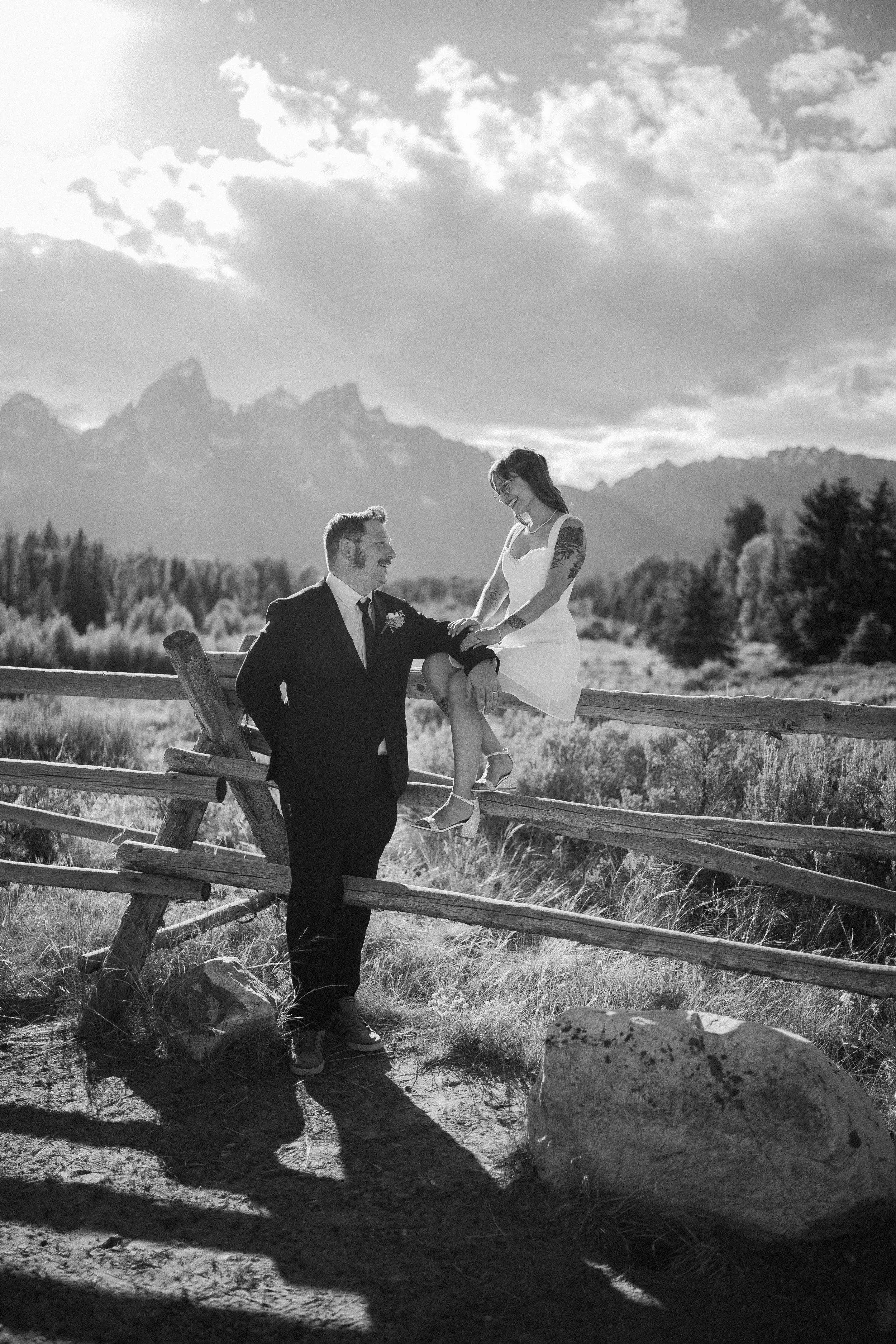 Black and white photo of a smiling bride in a short white dress sitting on a wooden fence beside her groom in a dark suit, with the Grand Teton mountains rising behind them in Wyoming.