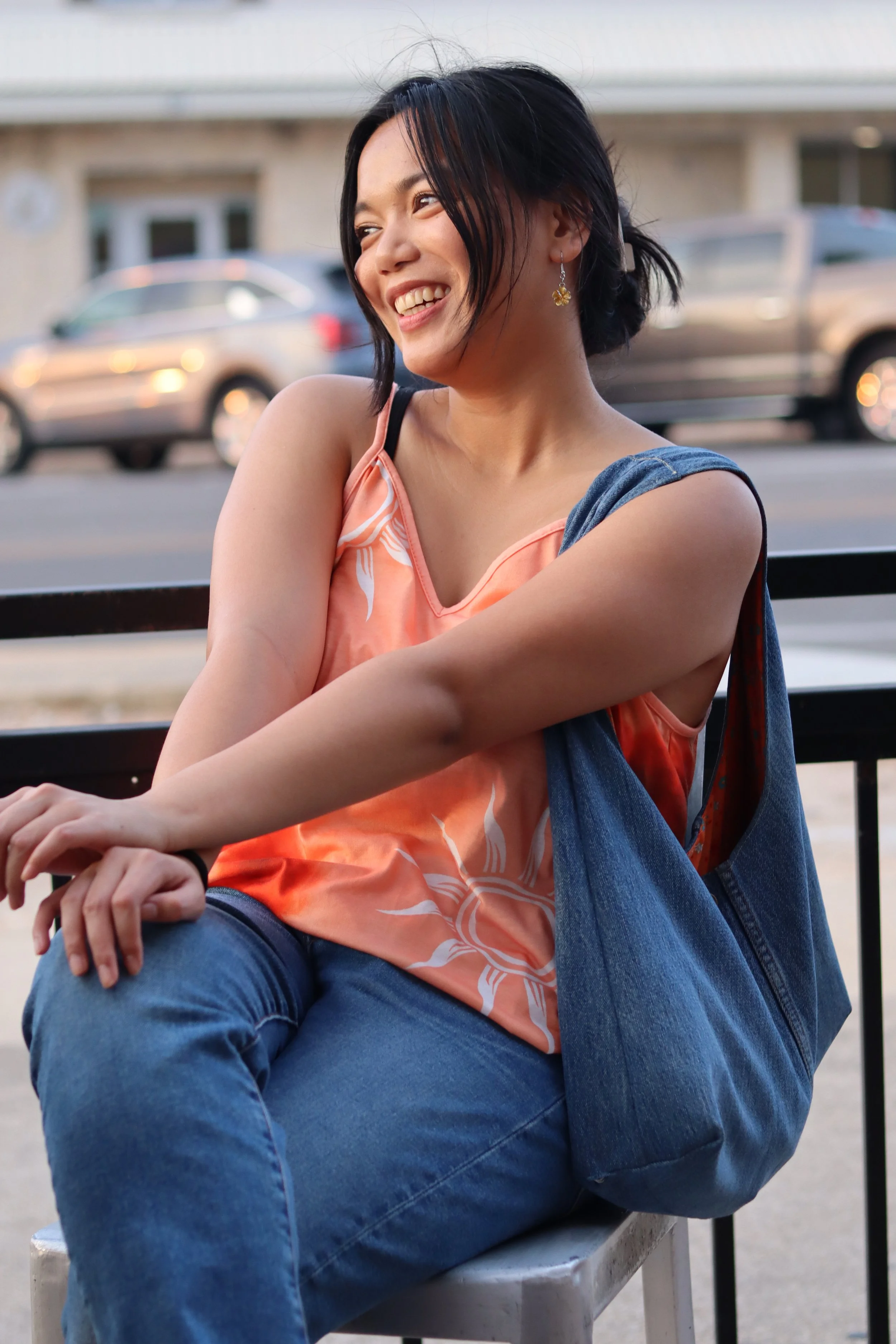 A woman sitting outdoors on a bench, smiling and looking to her left. She has short black hair and is wearing a pink and orange sleeveless top with white pattern, blue jeans, and flower-shaped earrings. There are cars and a building in the background