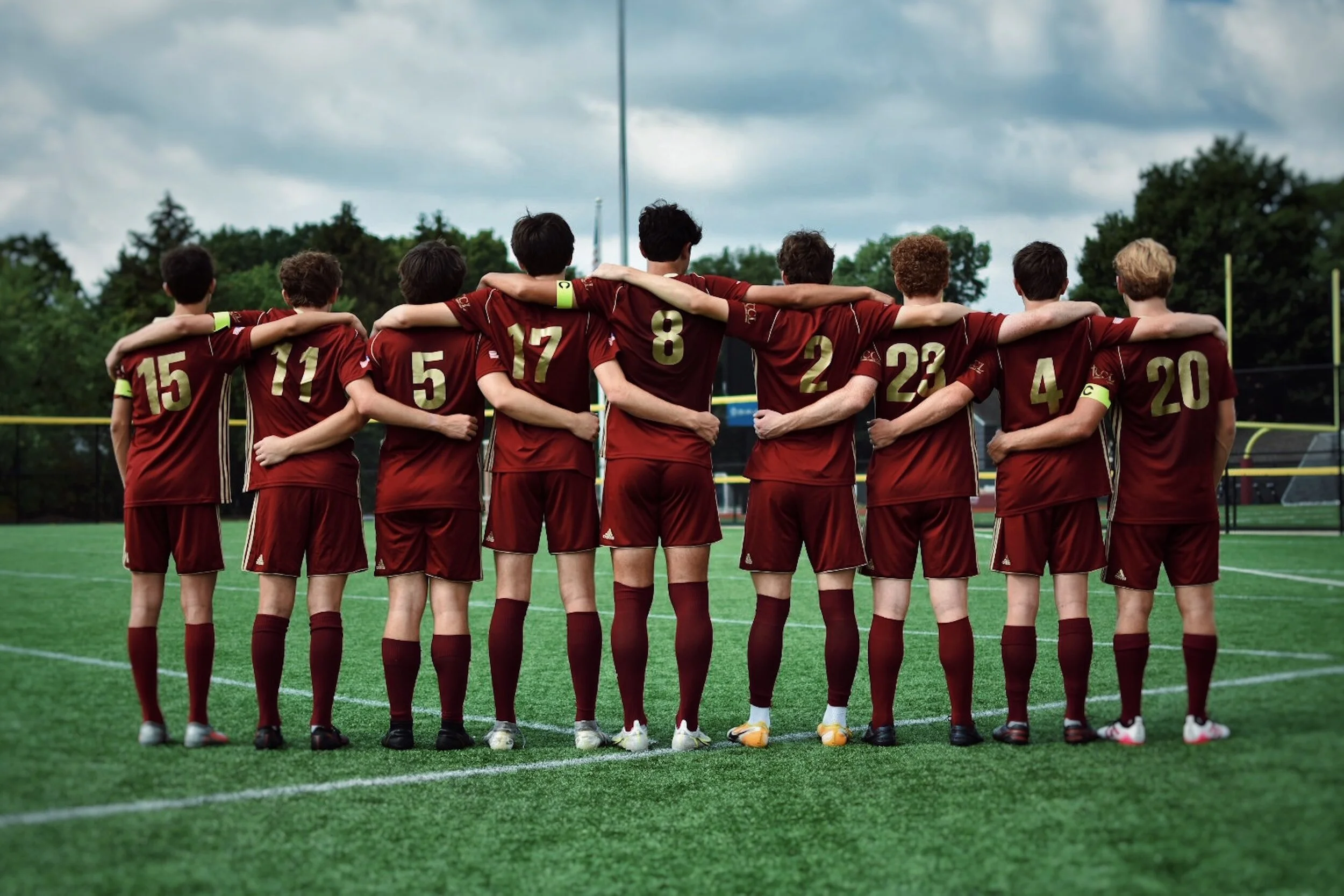 A youth football team stands together on a field with their arms around each other's shoulders, facing away from the camera, dressed in maroon uniforms with gold numbers, under a cloudy sky.