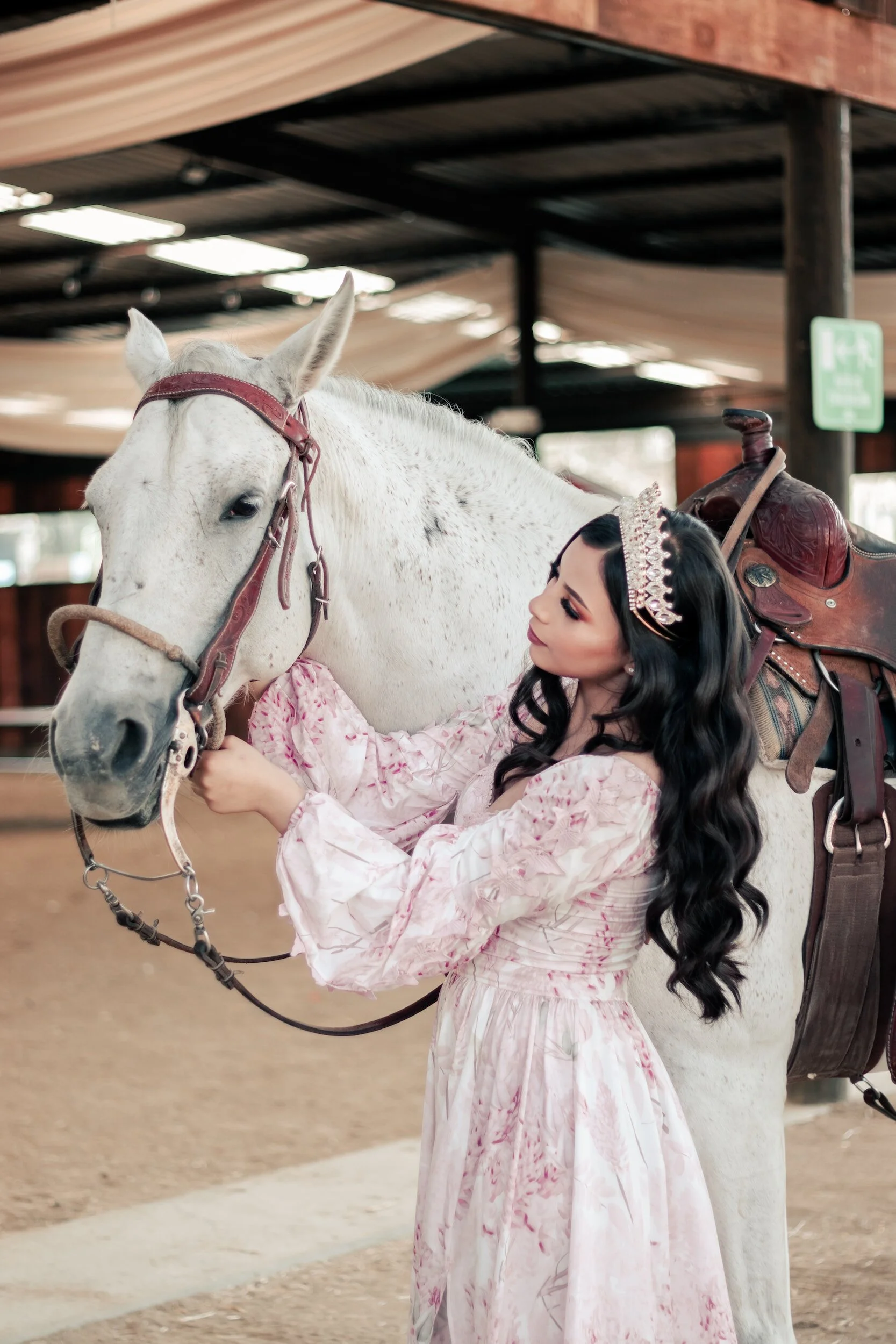 A woman with long black hair, wearing a pink dress and a tiara, is holding the bridle of a white horse with speckled gray markings inside a wooden structure. The woman is smiling and looking at the horse, which is saddled with a brown leather saddle.