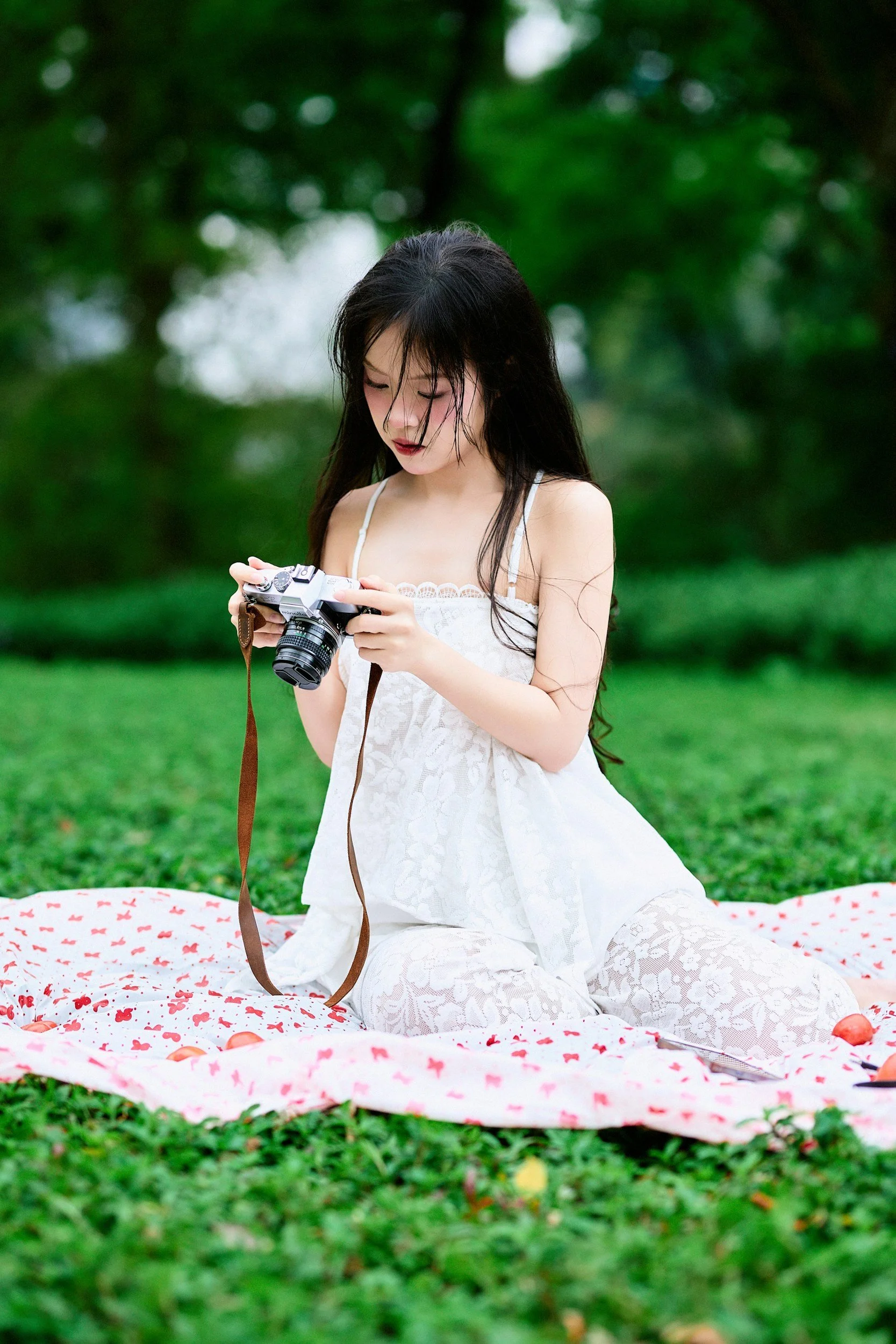 A young woman with long black hair, wearing a white lace dress, kneeling on a pink blanket with red bows, outdoors in a grassy area with trees in the background, holding a vintage film camera.