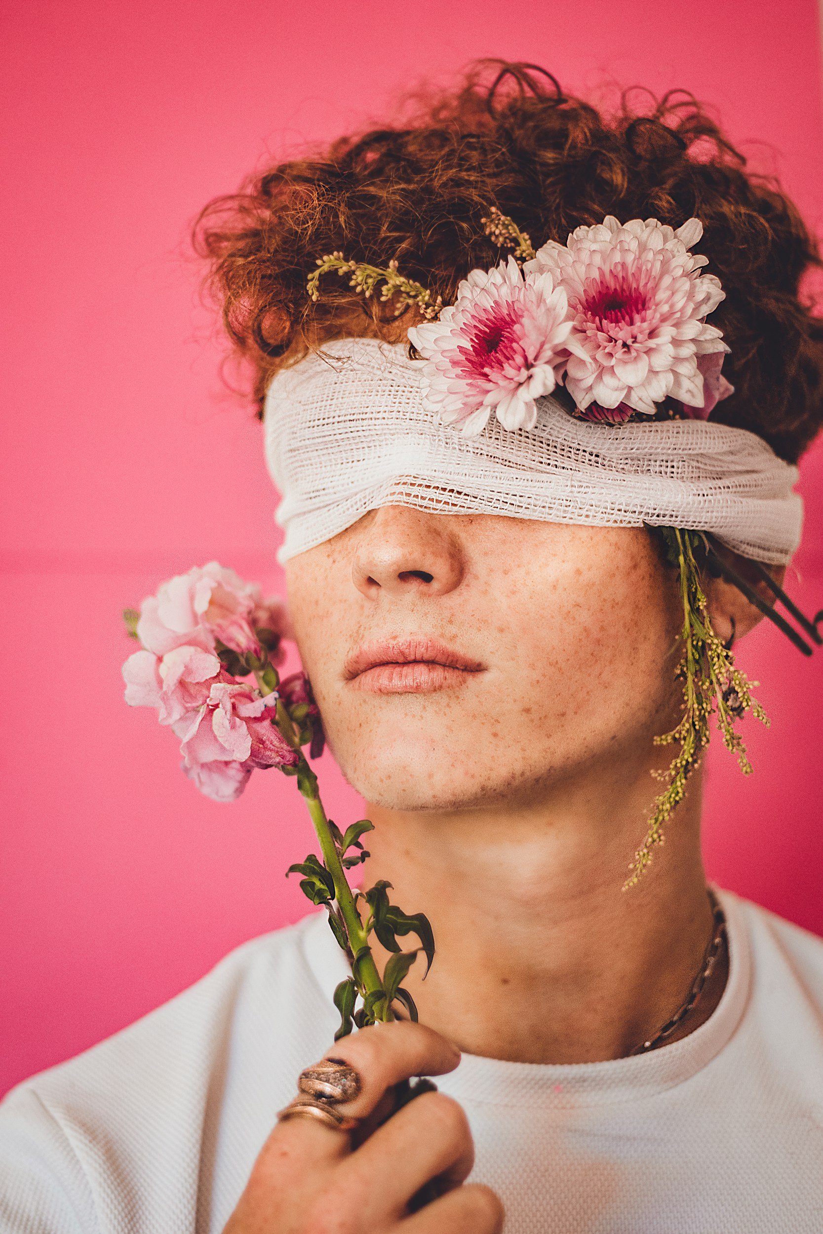 Person with curly hair has their eyes covered with a bandage and a large pink and white flower on their forehead, holding a pink flower stem, against a pink background.