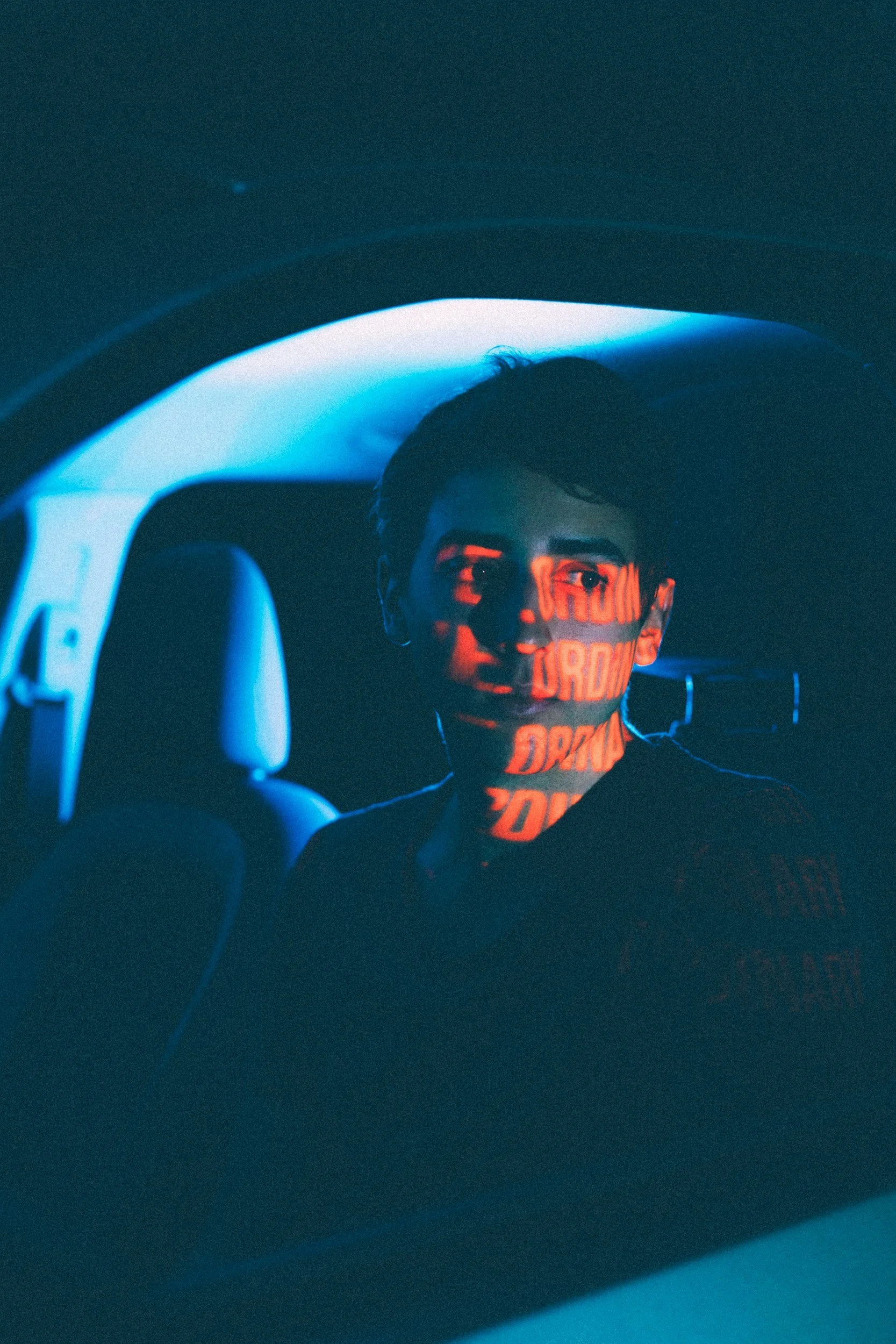 A young man with dark hair and face mask illuminated with red light, sitting inside a car at night, with the car's interior and window visible in the image.