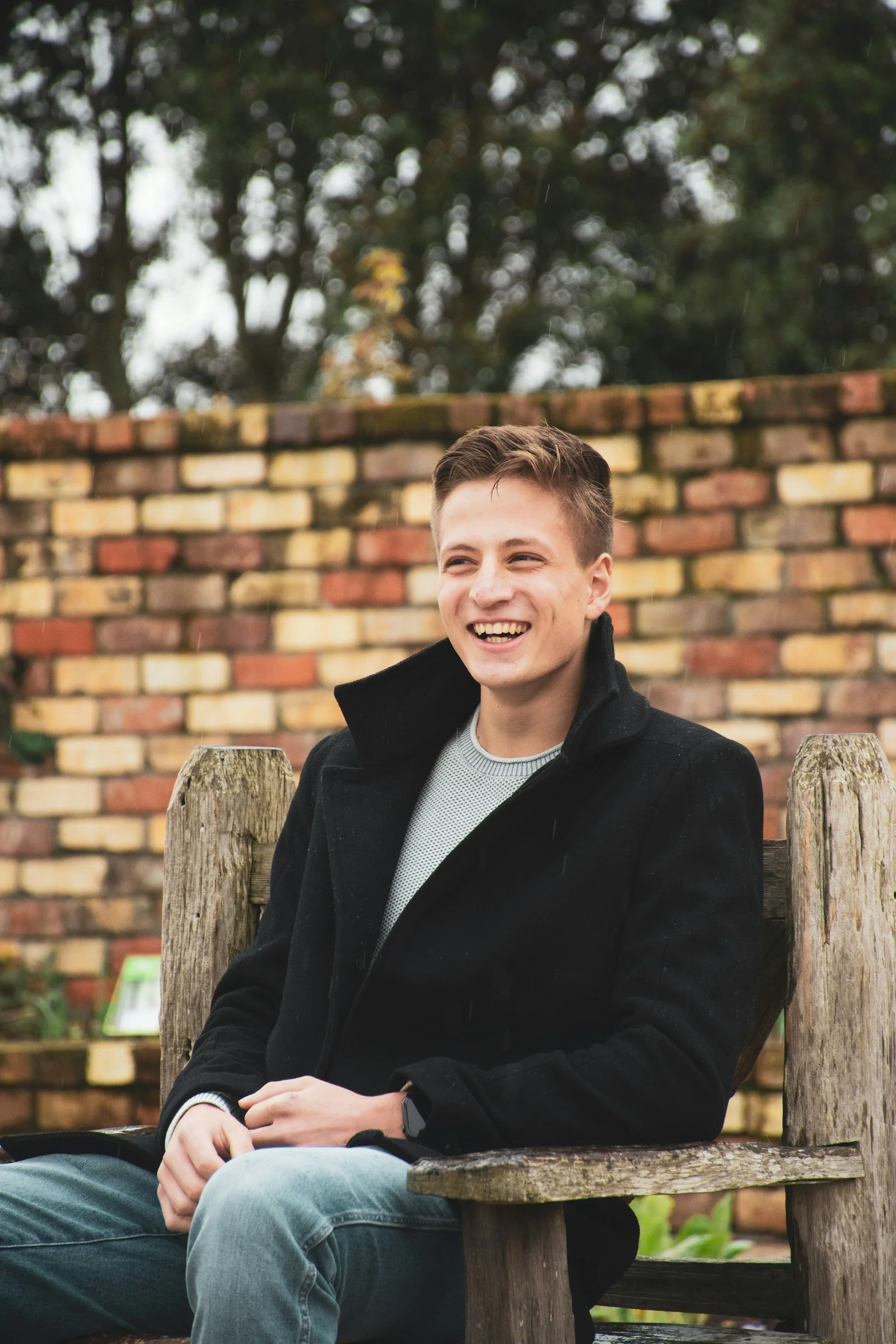 A young man with short brown hair, wearing a black jacket and a grey sweater, sitting on a wooden bench outdoors, smiling and holding a glass of wine. There is a brick wall behind him and trees with blurred leaves and sky in the background.