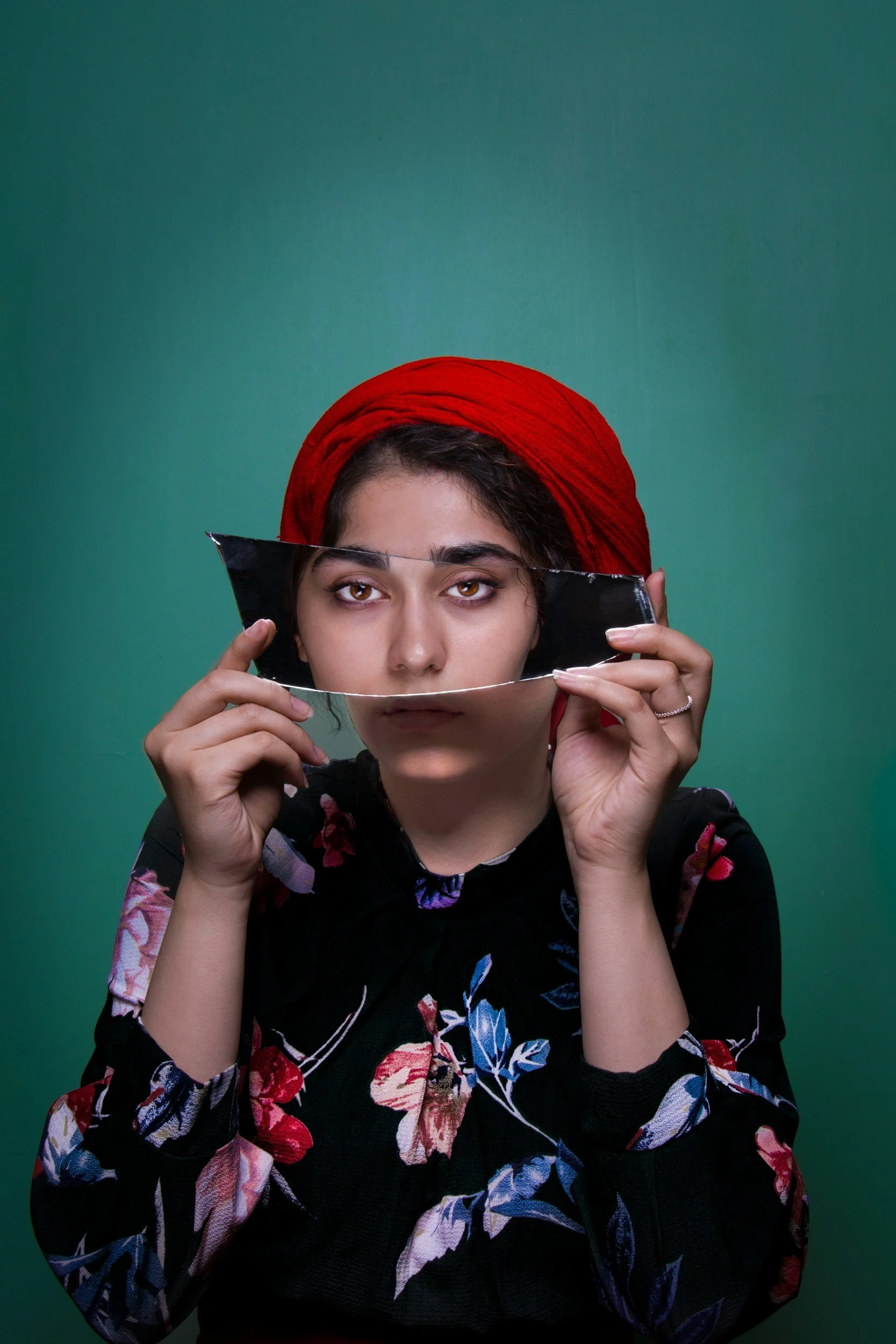 Portrait of a woman with dark hair, wearing a red headscarf and a black floral top, holding a broken mirror in front of her face against a green background.