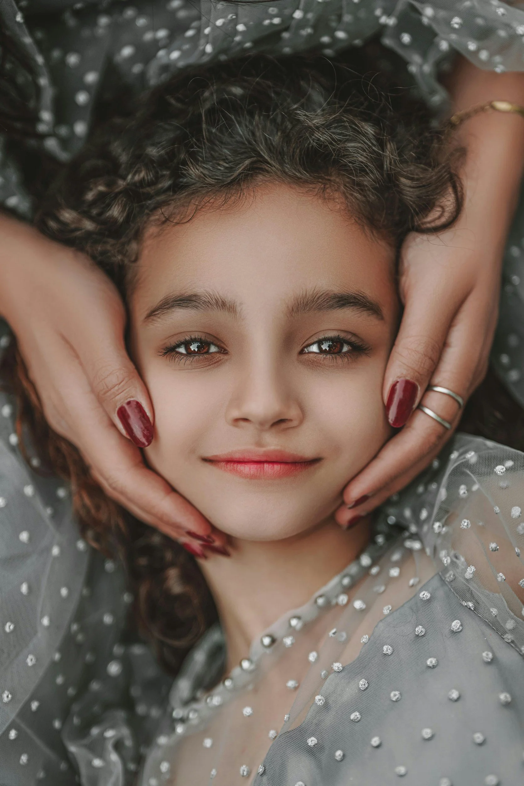 A young woman with curly hair lying down, holding her face with both hands, wearing a sheer, gray, polka-dot dress, and looking directly at the camera with a gentle smile.
