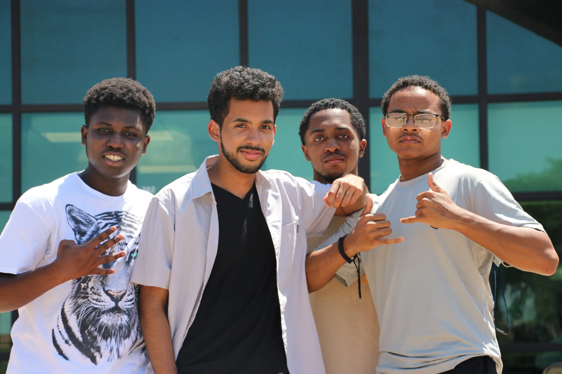 A group of four young men standing outside in front of a modern glass building, posing for the camera with confident expressions and making hand gestures.