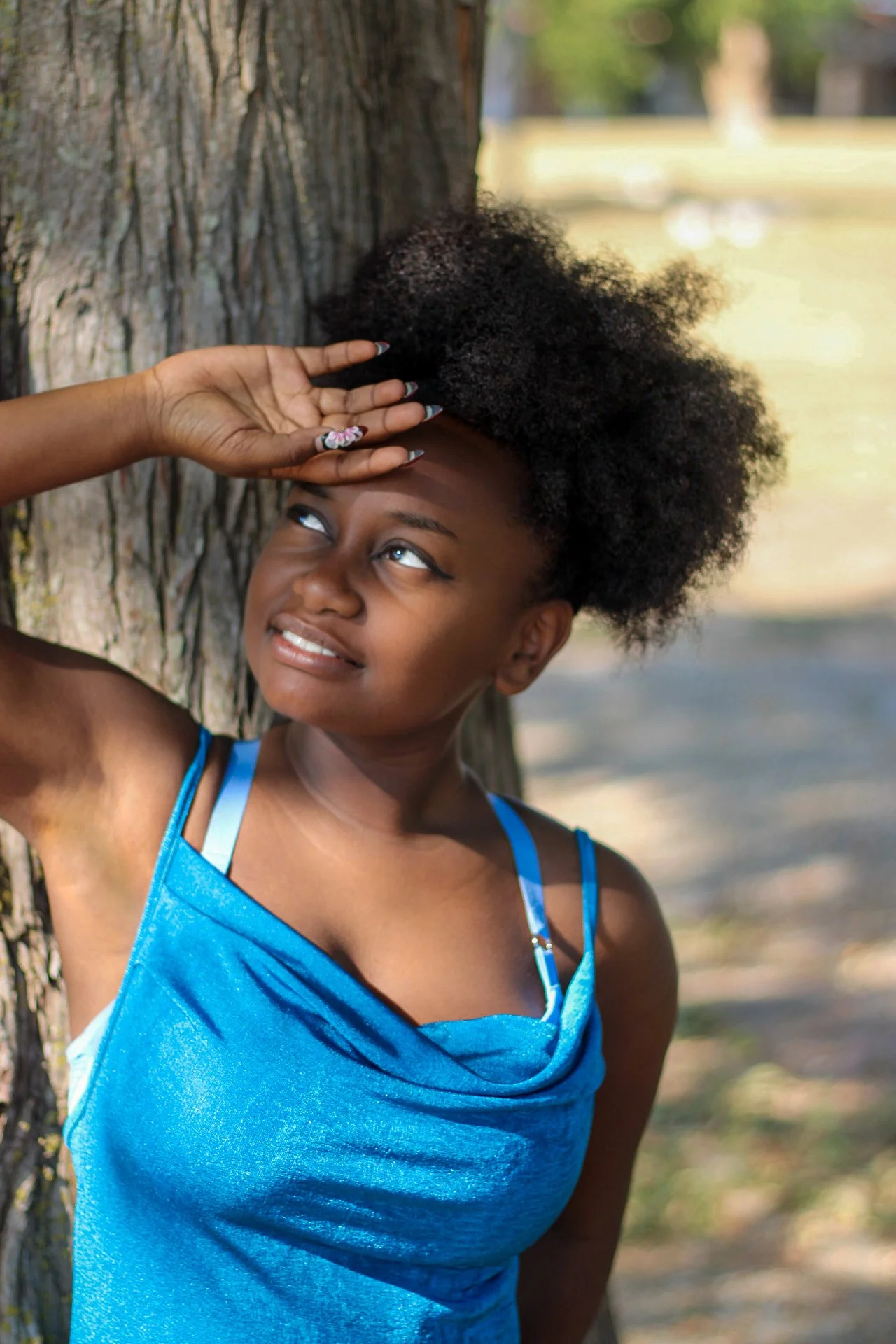 A young woman with natural curly hair wearing a blue dress, leaning against a tree outdoors, smiling and looking up, with a background of a park and sunlight filtering through trees.