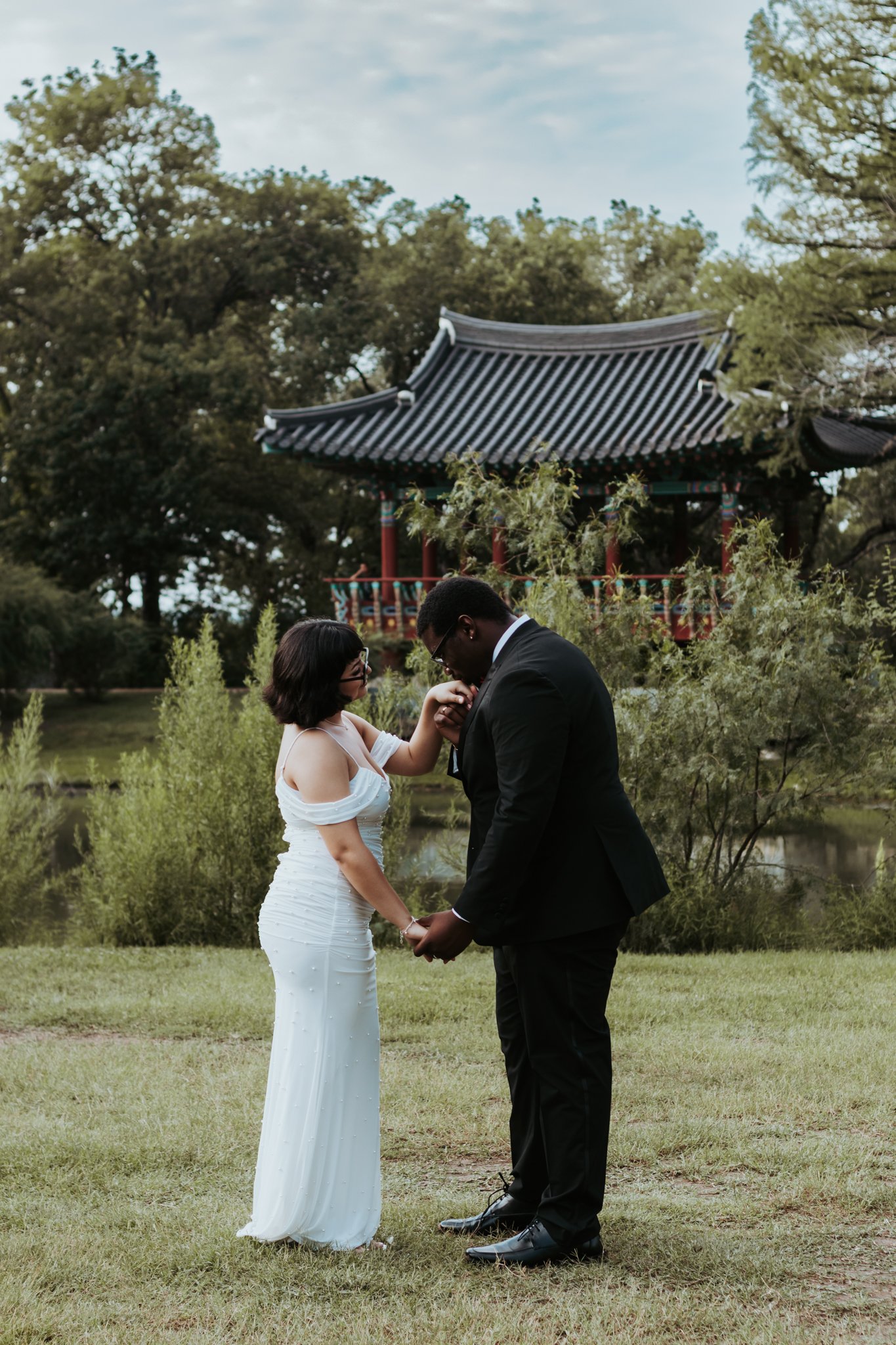 A bride and groom holding hands and kissing in a garden with a traditional Asian pavilion in the background.
