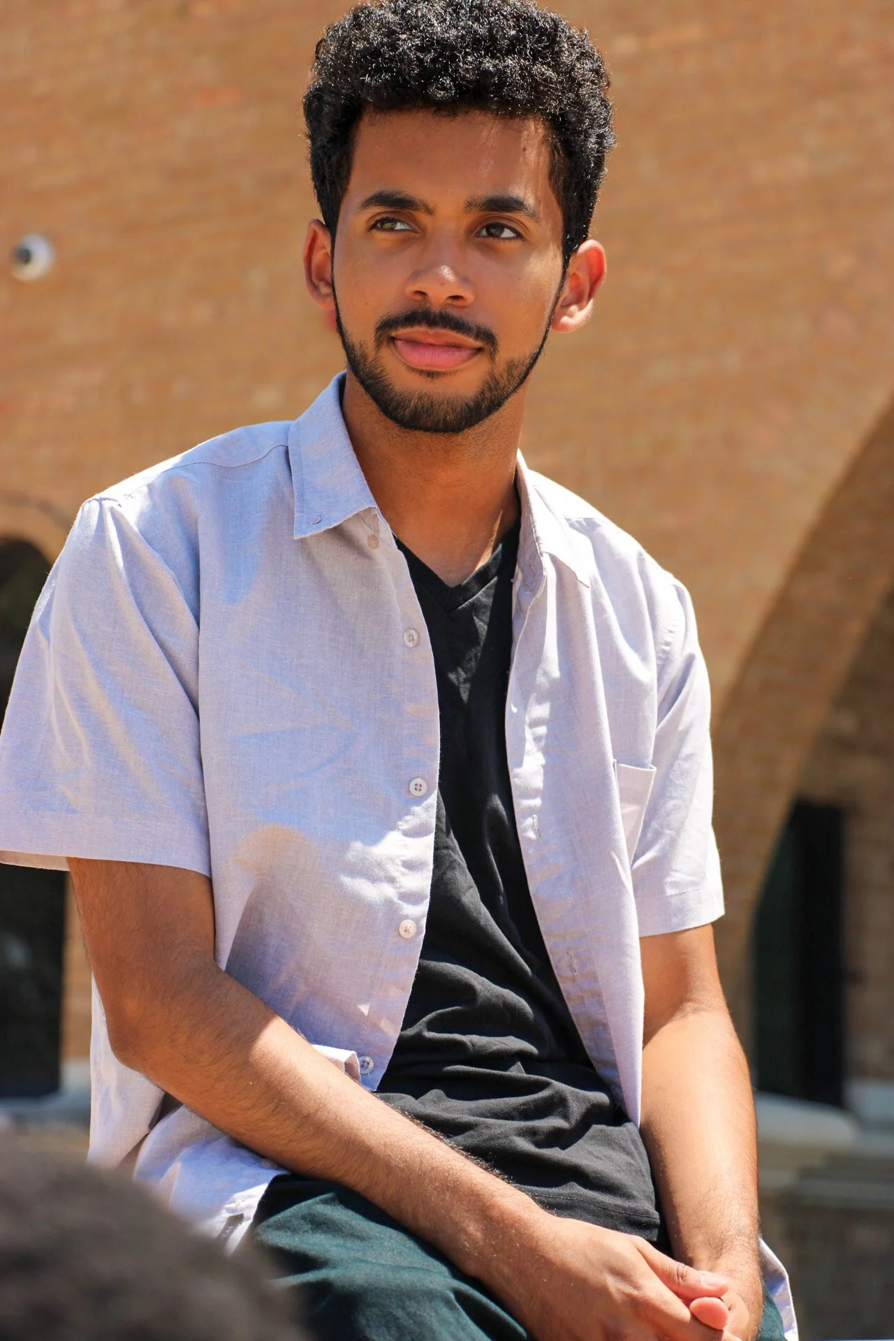 A young man with curly black hair and facial hair outdoors, wearing a light blue short-sleeved shirt over a black T-shirt, sitting on a bench with a brick building in the background.