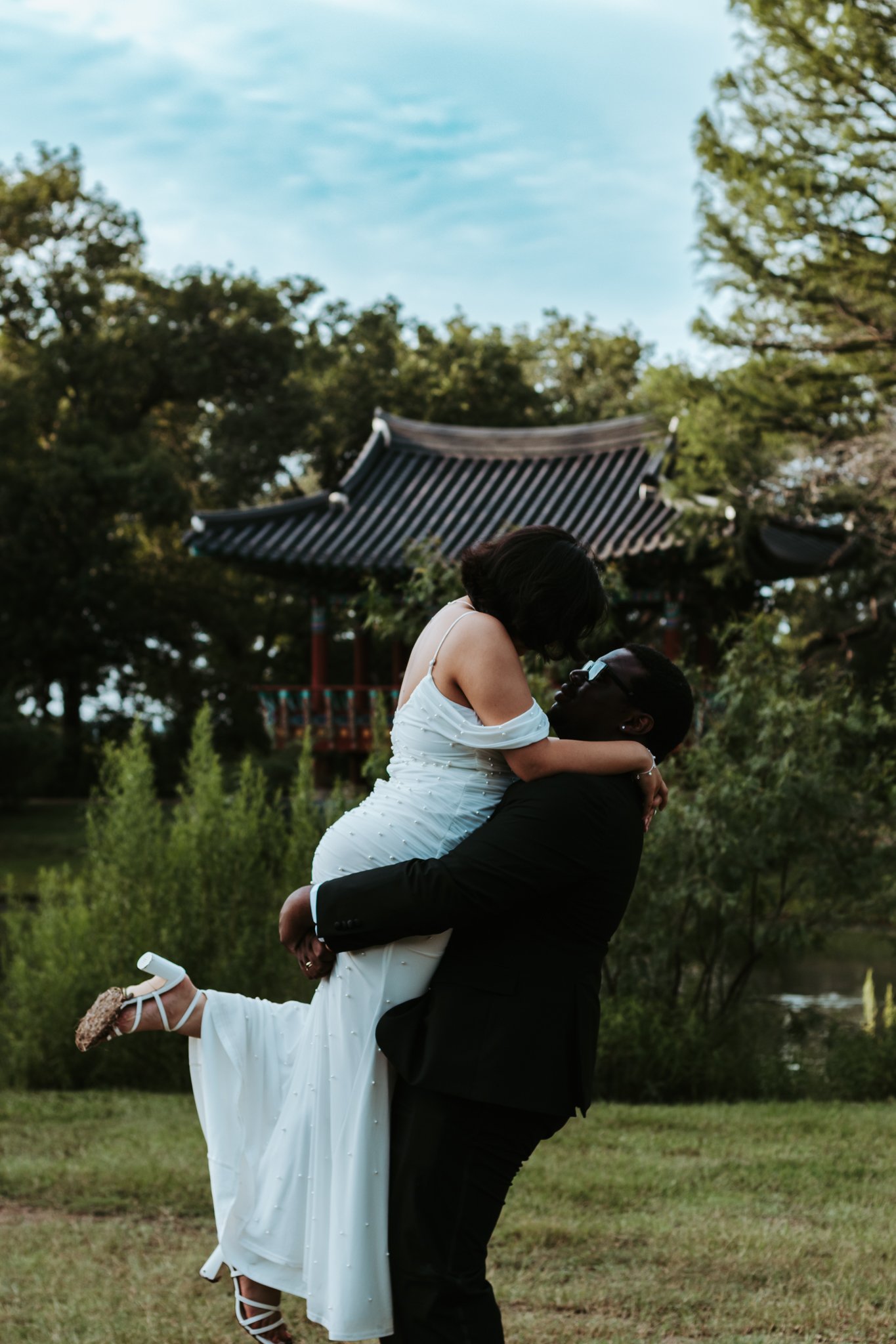 A couple is outdoors, with the man lifting and holding the woman as they embrace. The woman wears a white dress and shoes with a floral pattern, and the man wears a black suit. In the background, there are trees and a traditional Asian-style pavilion