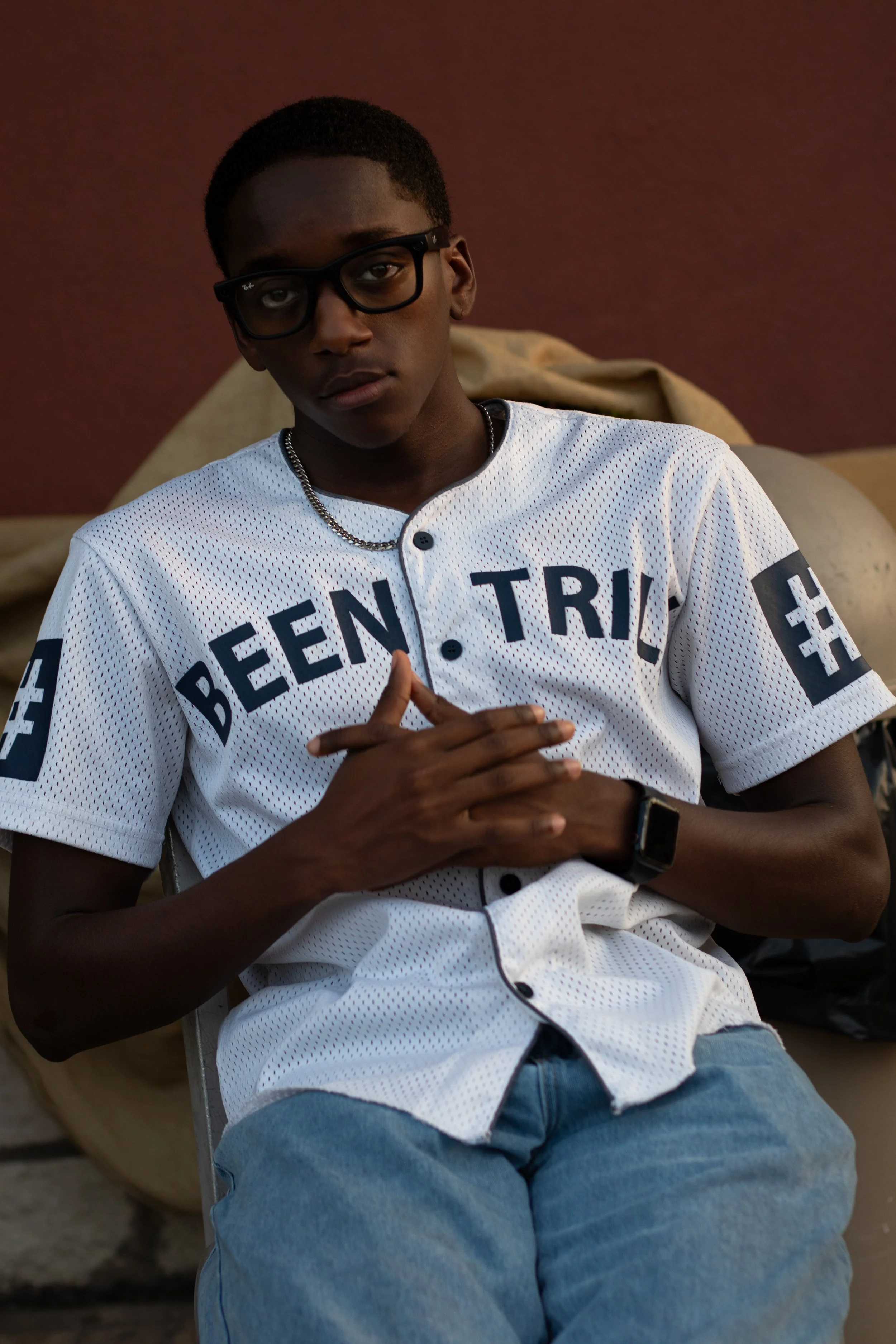 A young man with dark skin, short hair, wearing black glasses and a white athletic shirt with black lettering, sitting with his hands folded, against a dark background.