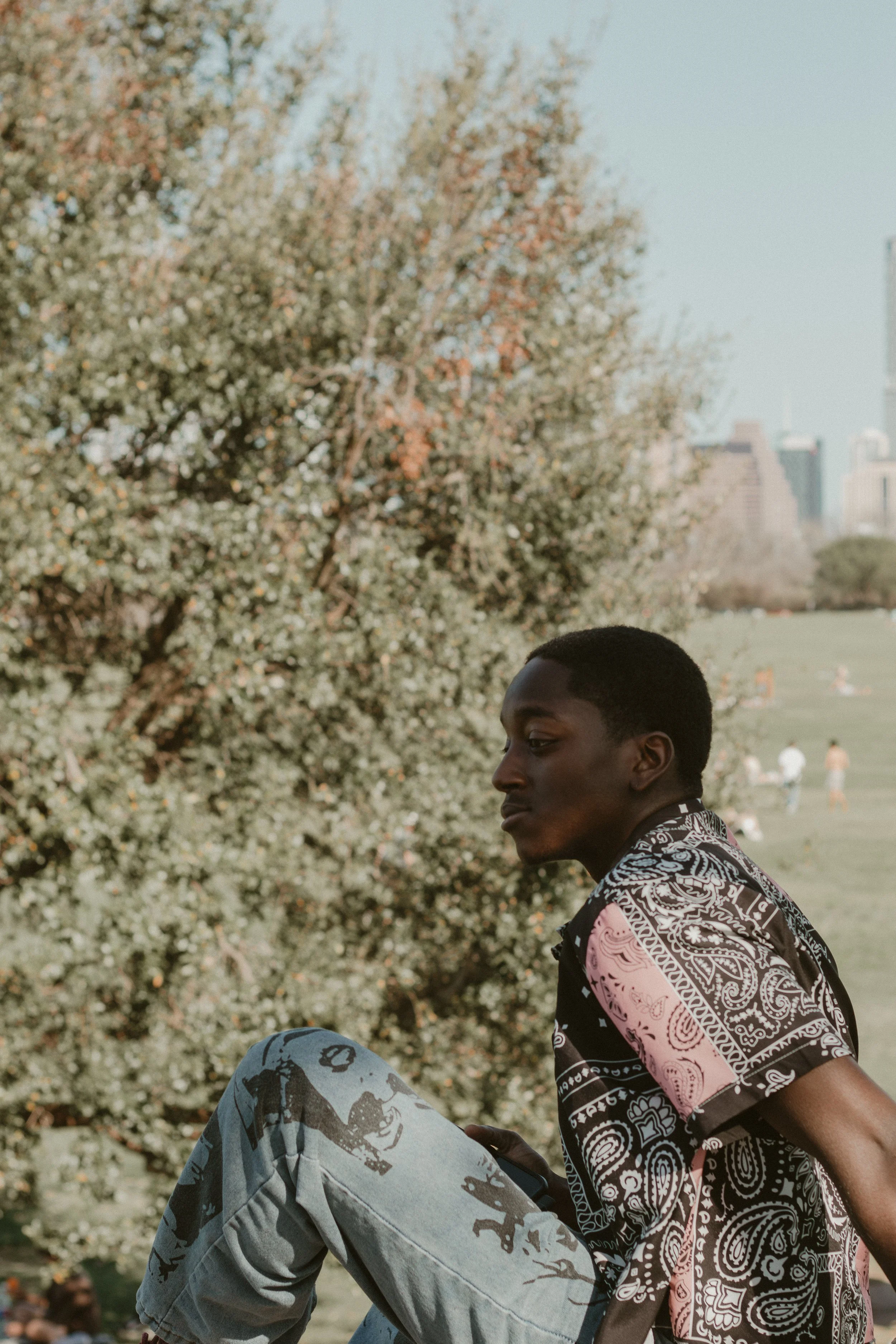 Side profile of a young man with dark skin and short hair, wearing a patterned short-sleeve shirt, sitting outdoors with a large tree and skyline in the background.