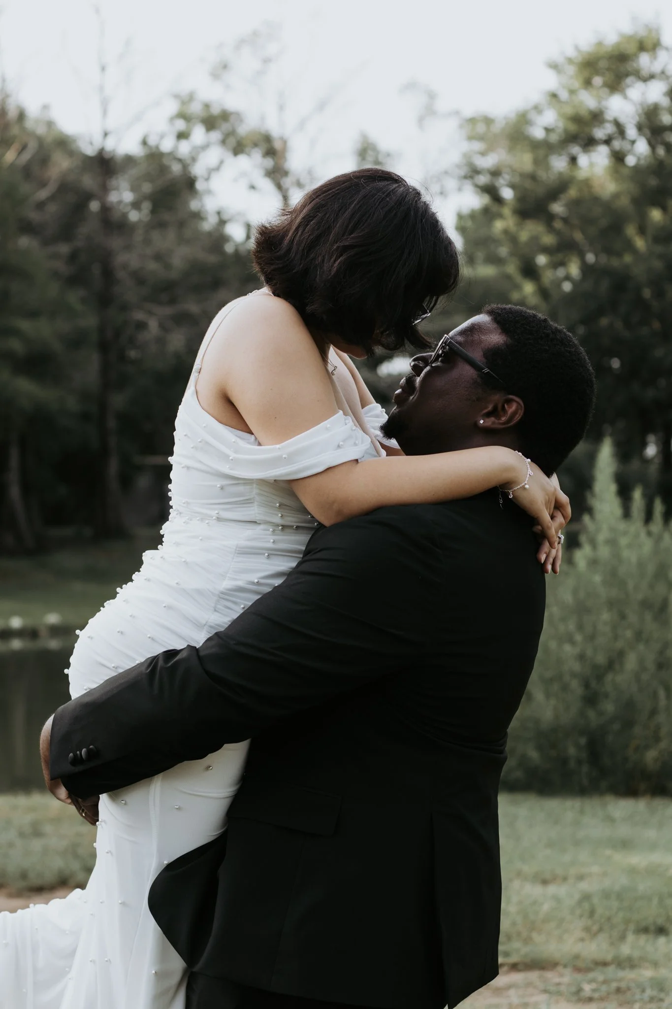 A couple outdoors during daytime, with the man lifting the woman in his arms. The woman is wearing a white dress and the man is in a black suit. Trees with green leaves are in the background.