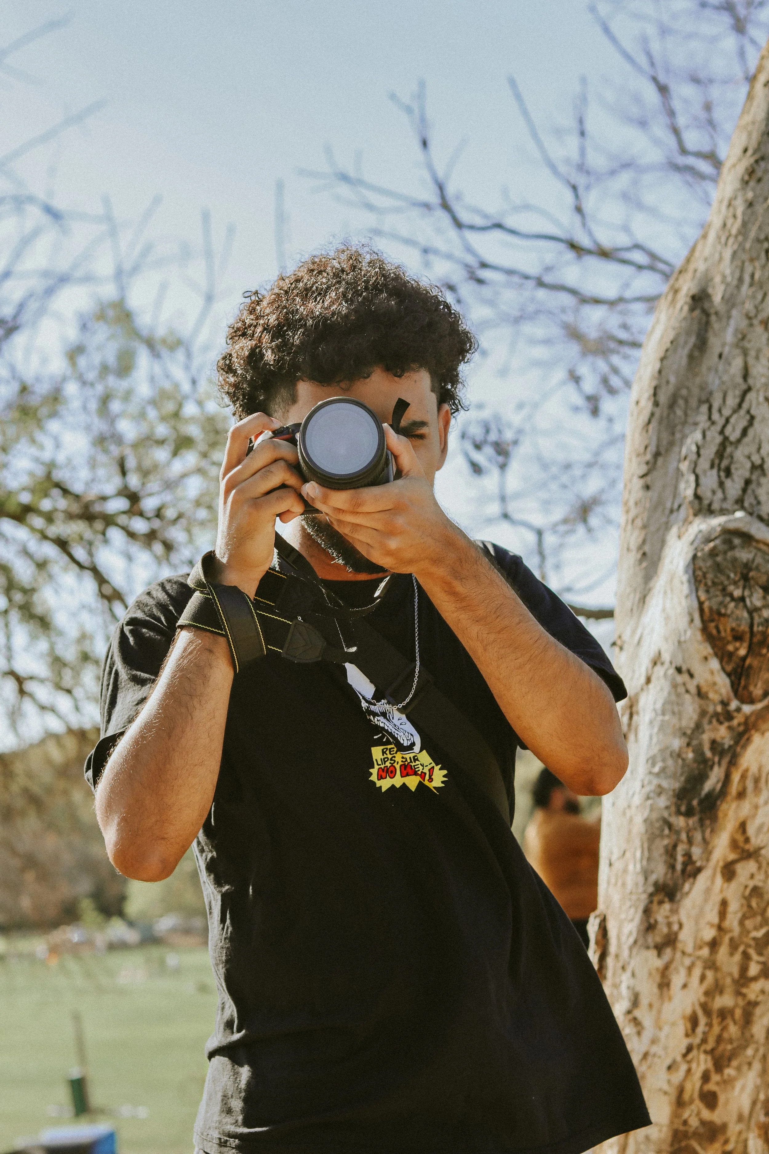 A man with curly hair taking a photo with a camera outdoors near a tree during daytime.