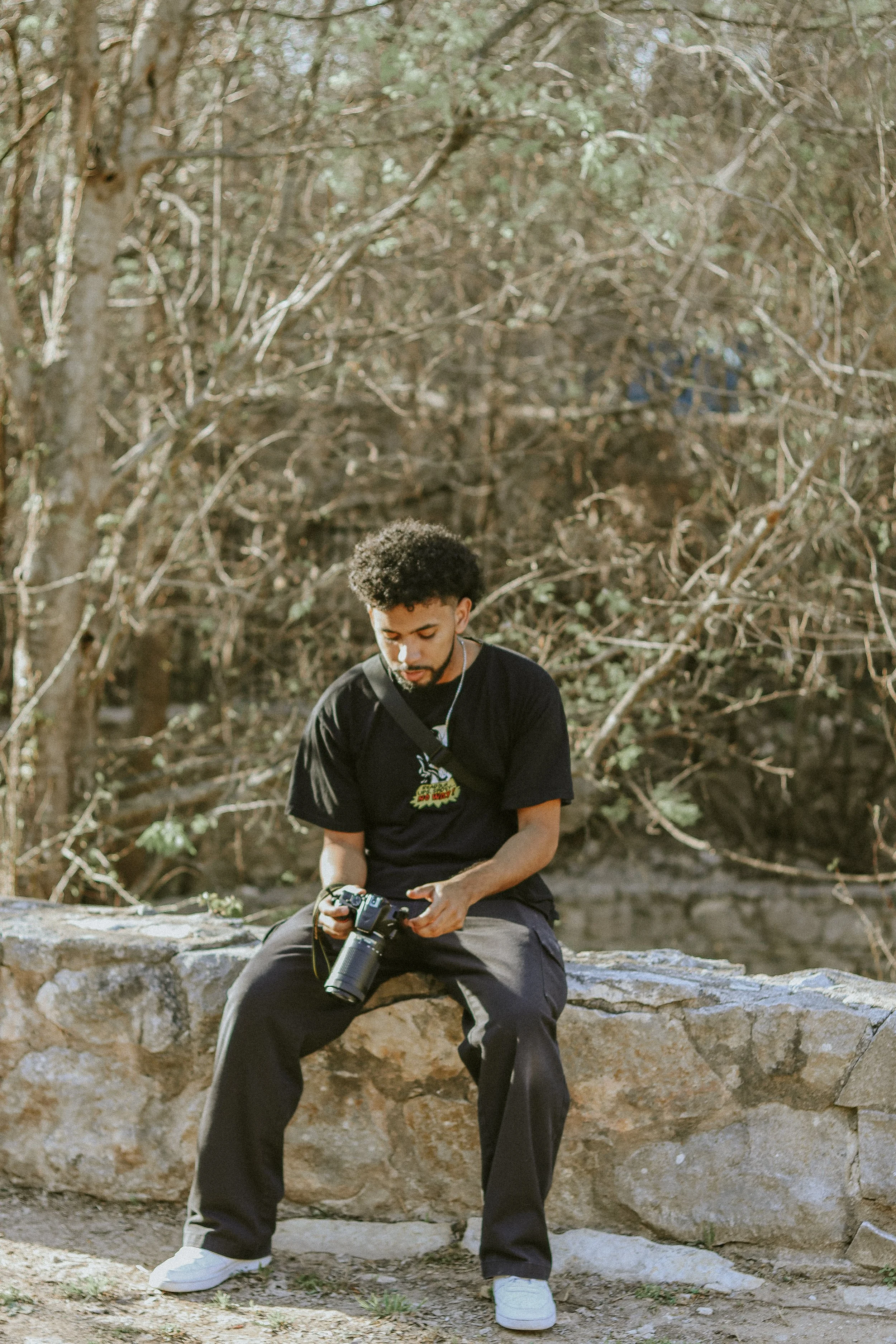 A young man with curly hair and a beard sitting on a large rock outdoors, holding a camera and looking down at it, with a background of trees and bushes.