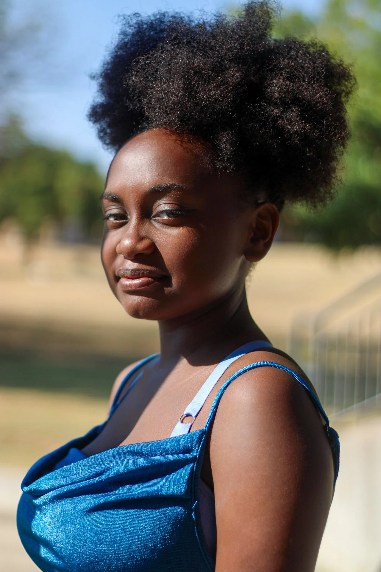 Young woman with dark skin and a large curly hairstyle outdoors, wearing a blue tank top.