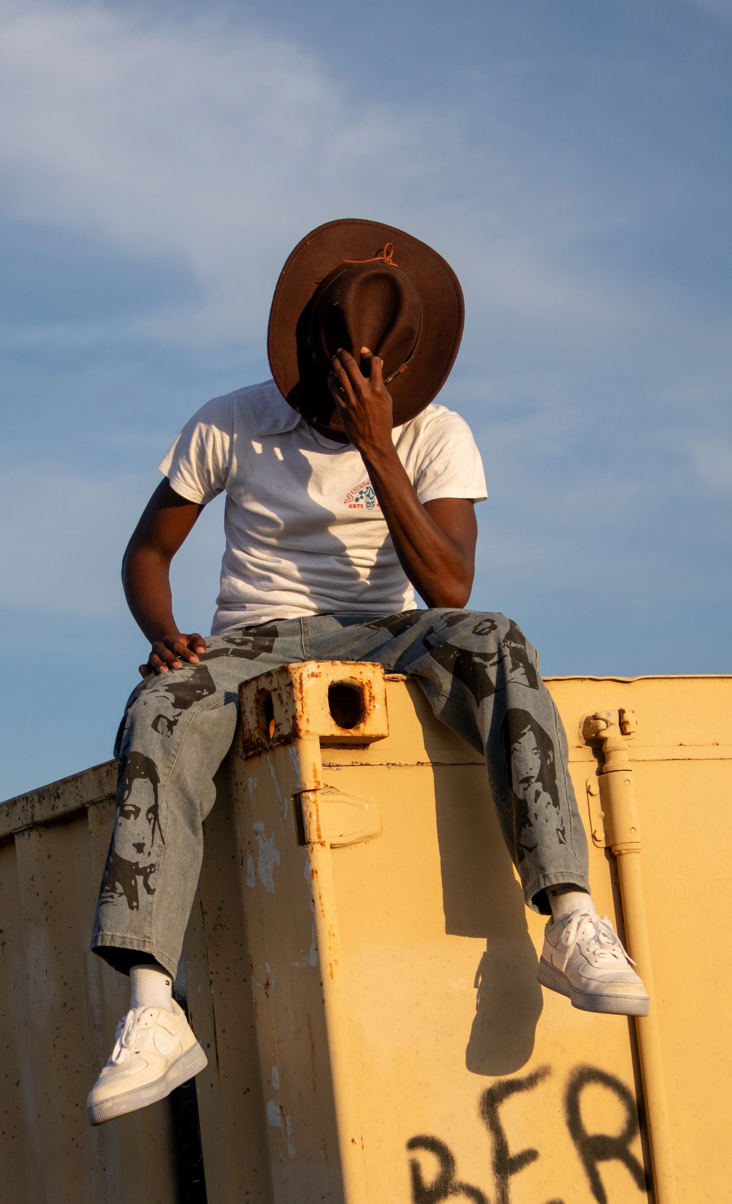 Person sitting on a yellow industrial structure, wearing a white t-shirt, gray camouflage pants, white sneakers, and a brown hat, with their face partially covered by their hand, against a blue sky with some clouds.