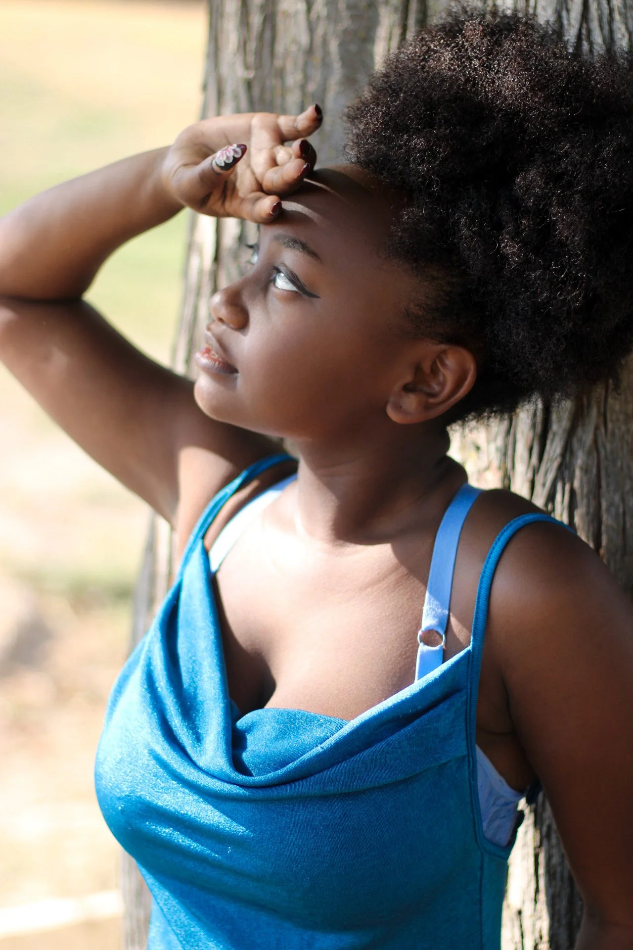 A young woman leaning against a tree outdoors, wearing a blue dress, with curly hair, looking thoughtfully into the distance.