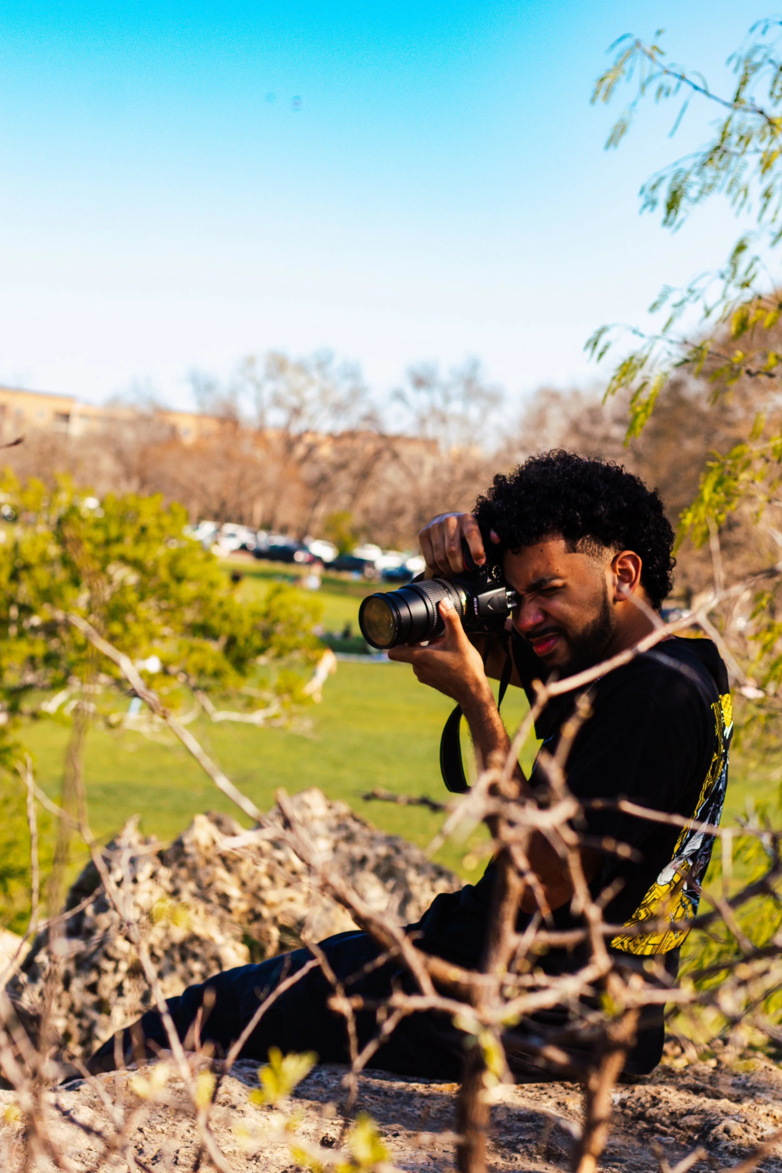 A young man with curly hair and a beard taking a photograph with a camera outdoors near some fallen branches and trees, with a park and parked cars in the background during daytime.