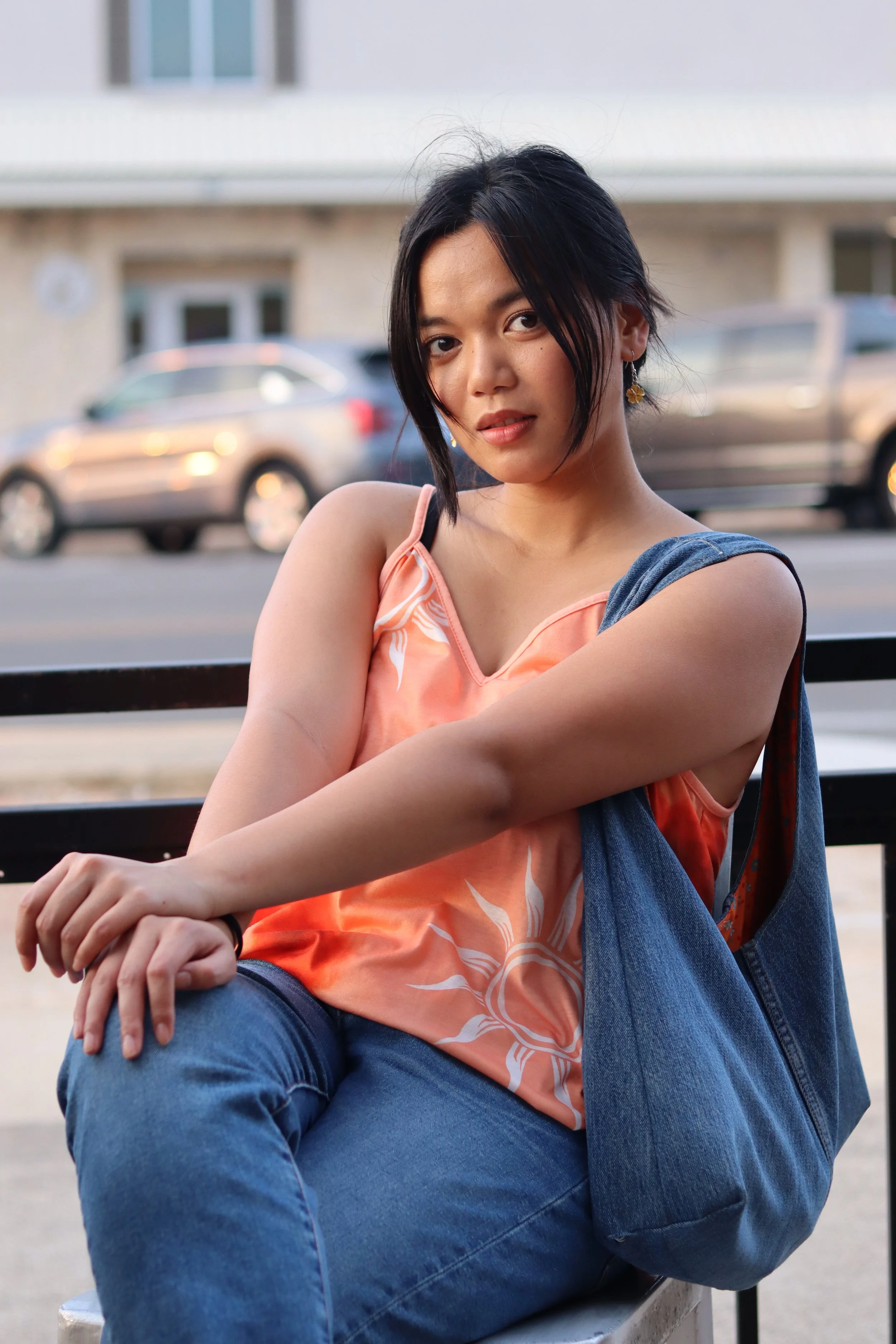 A young woman with short black hair, wearing a peach-colored top with white floral patterns and blue jeans, sitting on a bench outdoors. She has an earring in her left ear and is looking at the camera with a slight smile. The background features parked cars and a building.